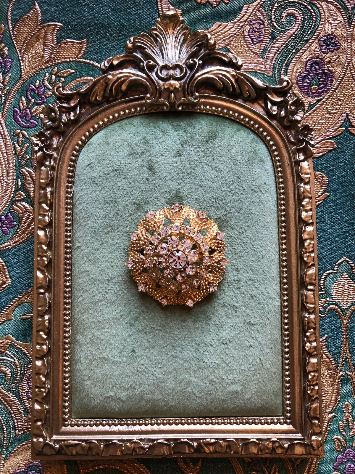 Broochella Caterina Vintage Gold Crystal Brooch featuring clear faceted crystals in an ornate textured gold-tone circular setting. It is photographed on a green velvet tray with an ornate gold frame. The background is green and gold damask wallpaper.