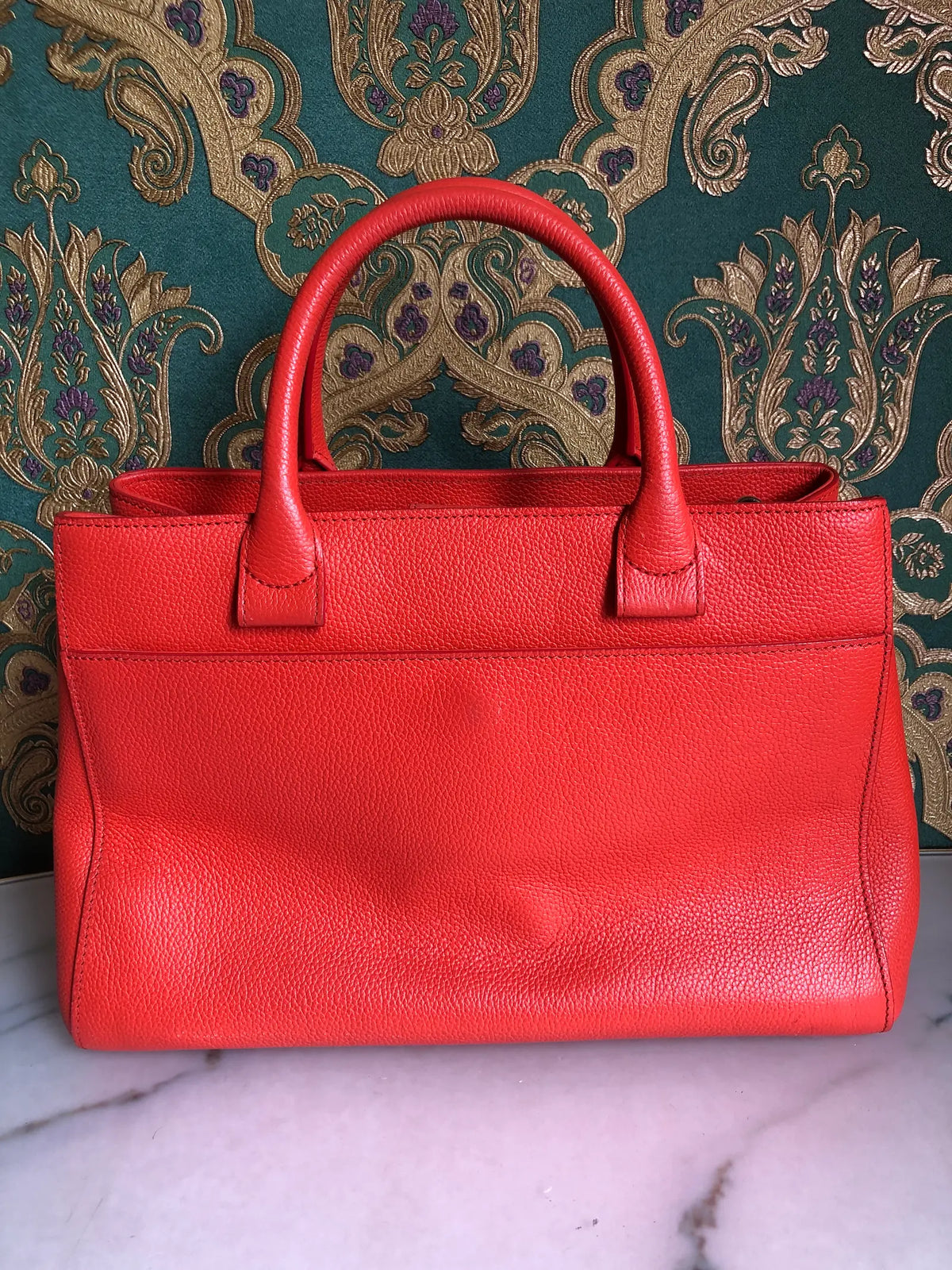 Broochella Pre-owned Chanel Executive Tote Bag in orange caviar leather with top handles and detachable shoulder strap. The background is green and gold damask wallpaper and a marble table