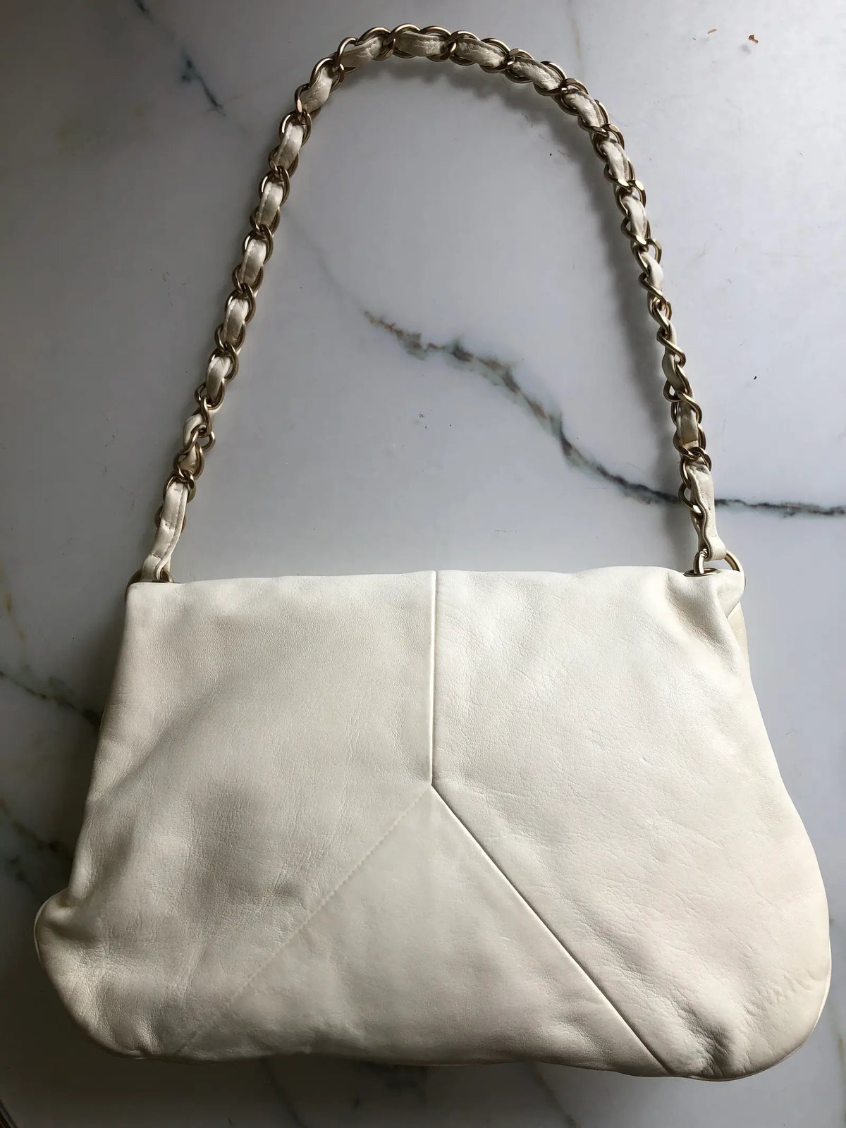 Broochella Chanel shoulder bag in ivory leather with a fold-over flap, gold-tone leather-woven chain strap and enamel CC ball charm. Small marks are visible on the front. Photographed against a white marble background.