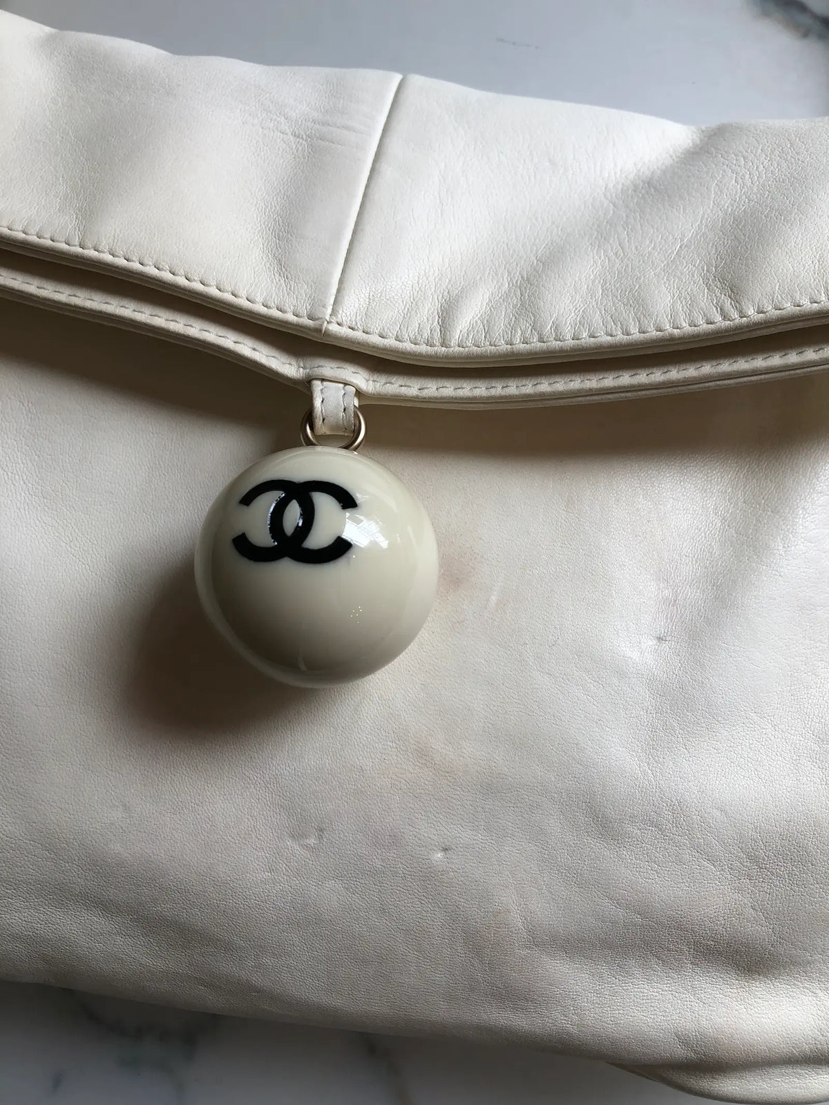 Broochella Chanel shoulder bag in ivory leather with a fold-over flap, gold-tone leather-woven chain strap and enamel CC ball charm. Small marks are visible on the front. Photographed against a white marble background.