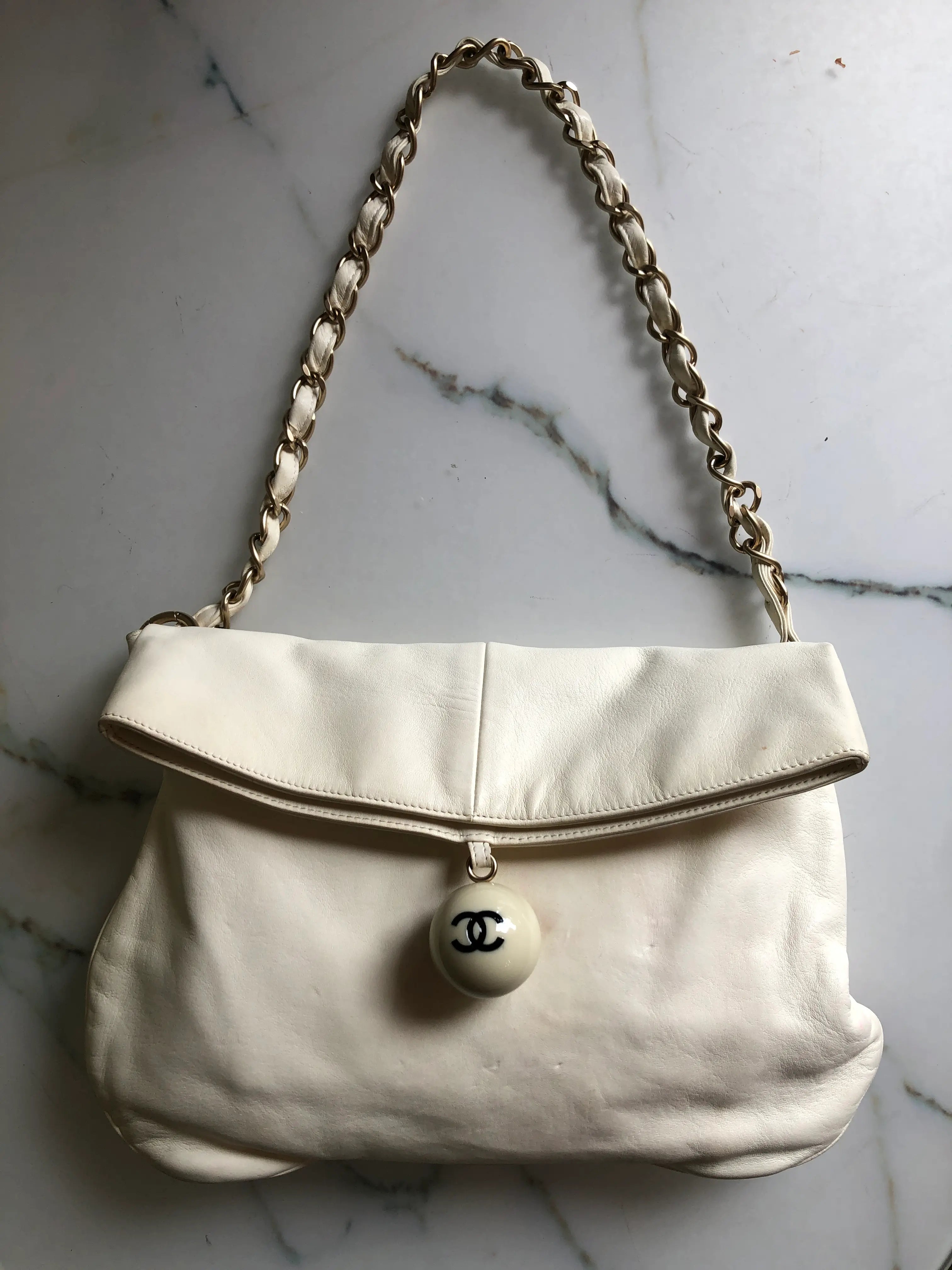Broochella Chanel shoulder bag in ivory leather with a fold-over flap, gold-tone leather-woven chain strap and enamel CC ball charm. Small marks are visible on the front. Photographed against a white marble background.