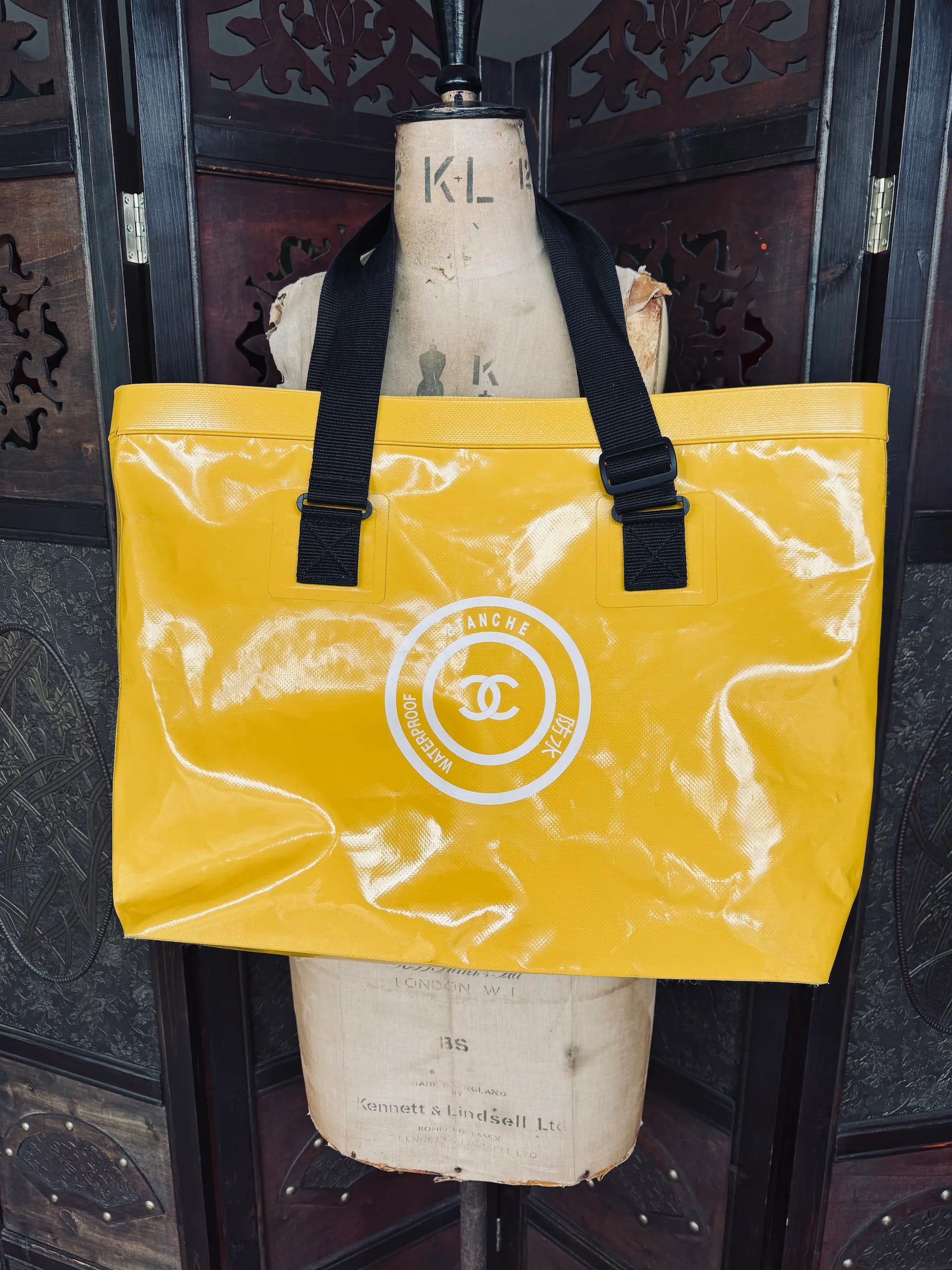 Broochella authentic designer Chanel large yellow waterproof tote bag with thick black straps. It is hanging over a vintage mannequin. The background is an ornate dark wood room divider
