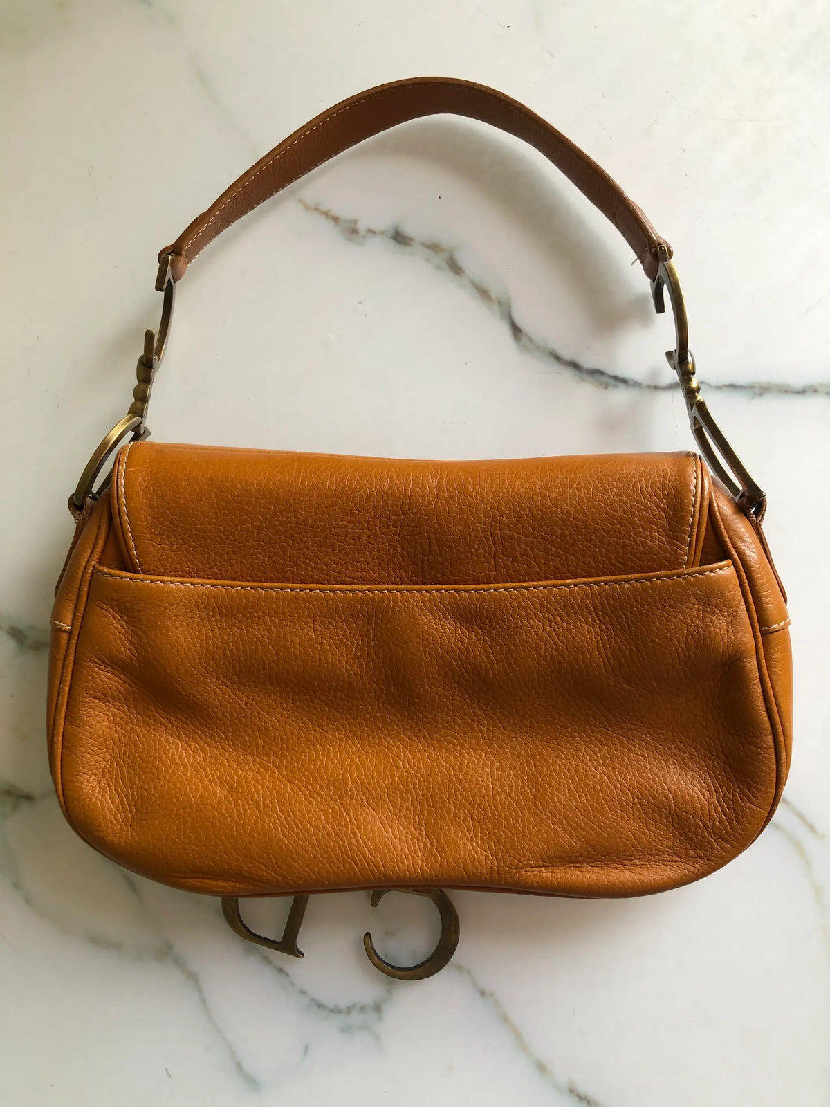The back view of Broochella Christian Dior vintage 2002 Saddle Bag in tan grained calfskin leather with antique gold-tone CD hardware and hanging D charm. Pictured on a white marble surface.
