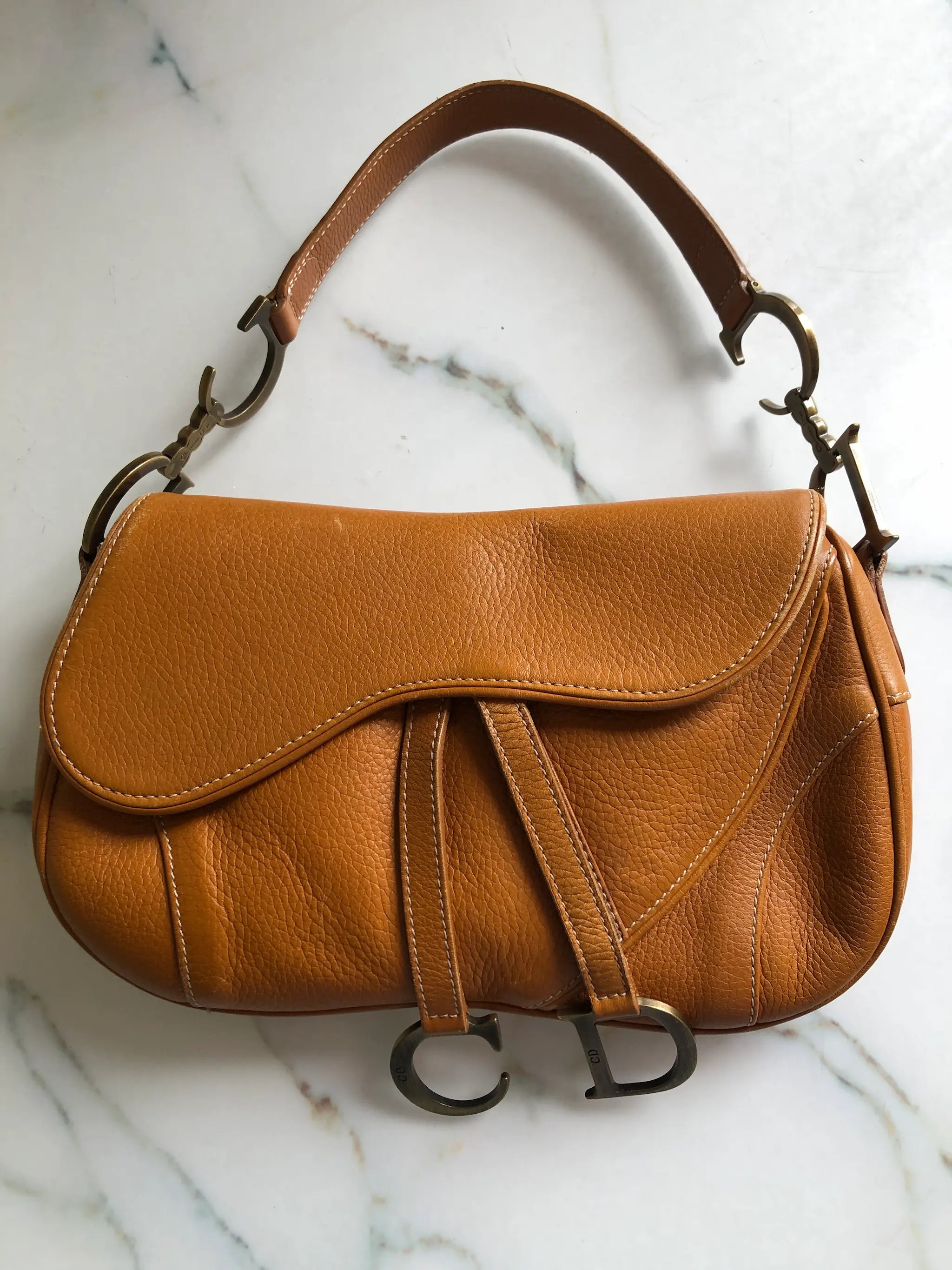 Broochella Christian Dior vintage 2002 Saddle Bag in tan grained calfskin leather with antique gold-tone CD hardware and hanging D charm. Pictured on a white marble surface.