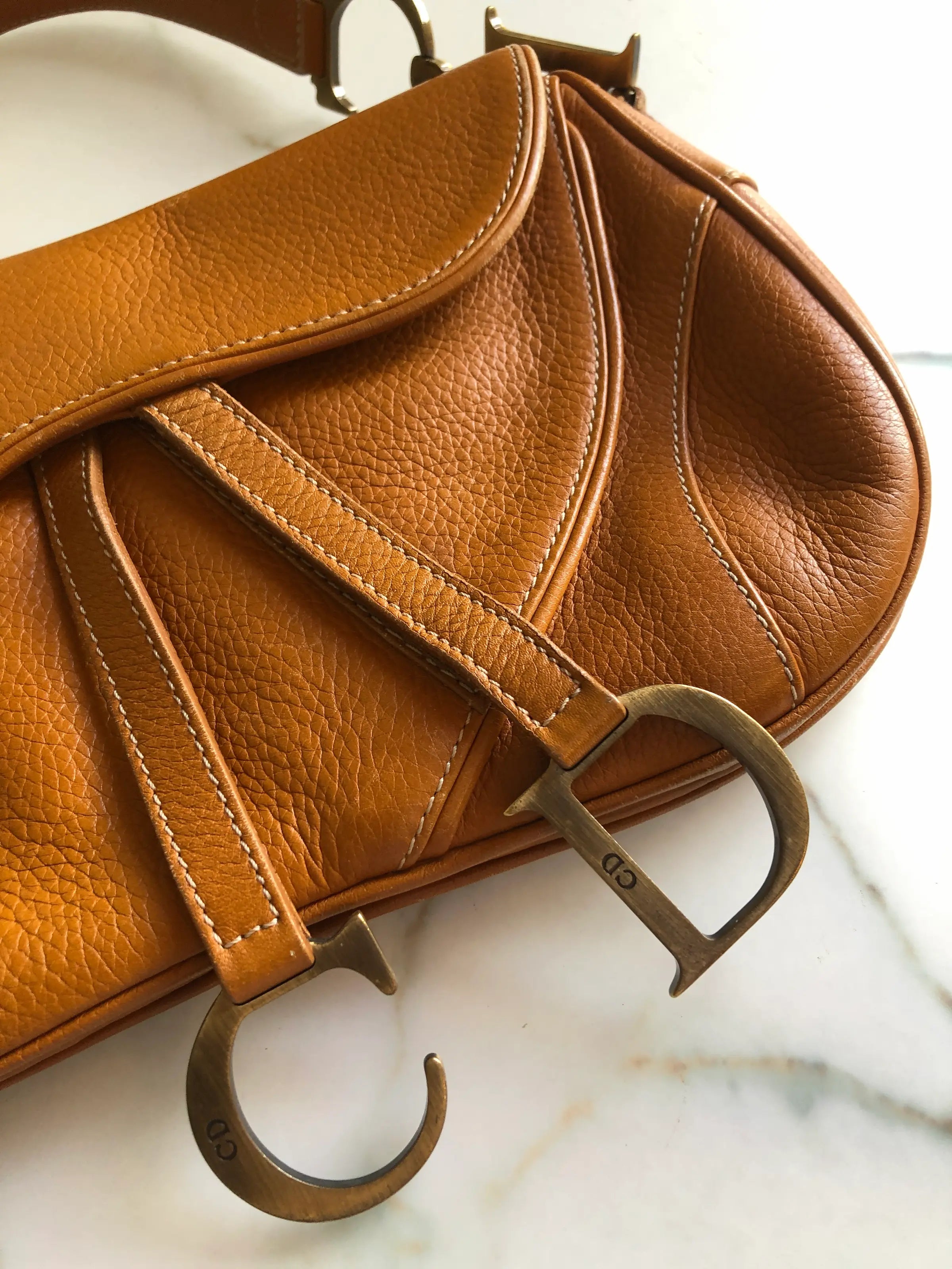 Broochella Christian Dior vintage 2002 Saddle Bag in tan grained calfskin leather with antique gold-tone CD hardware and hanging D charm. Pictured on a white marble surface.