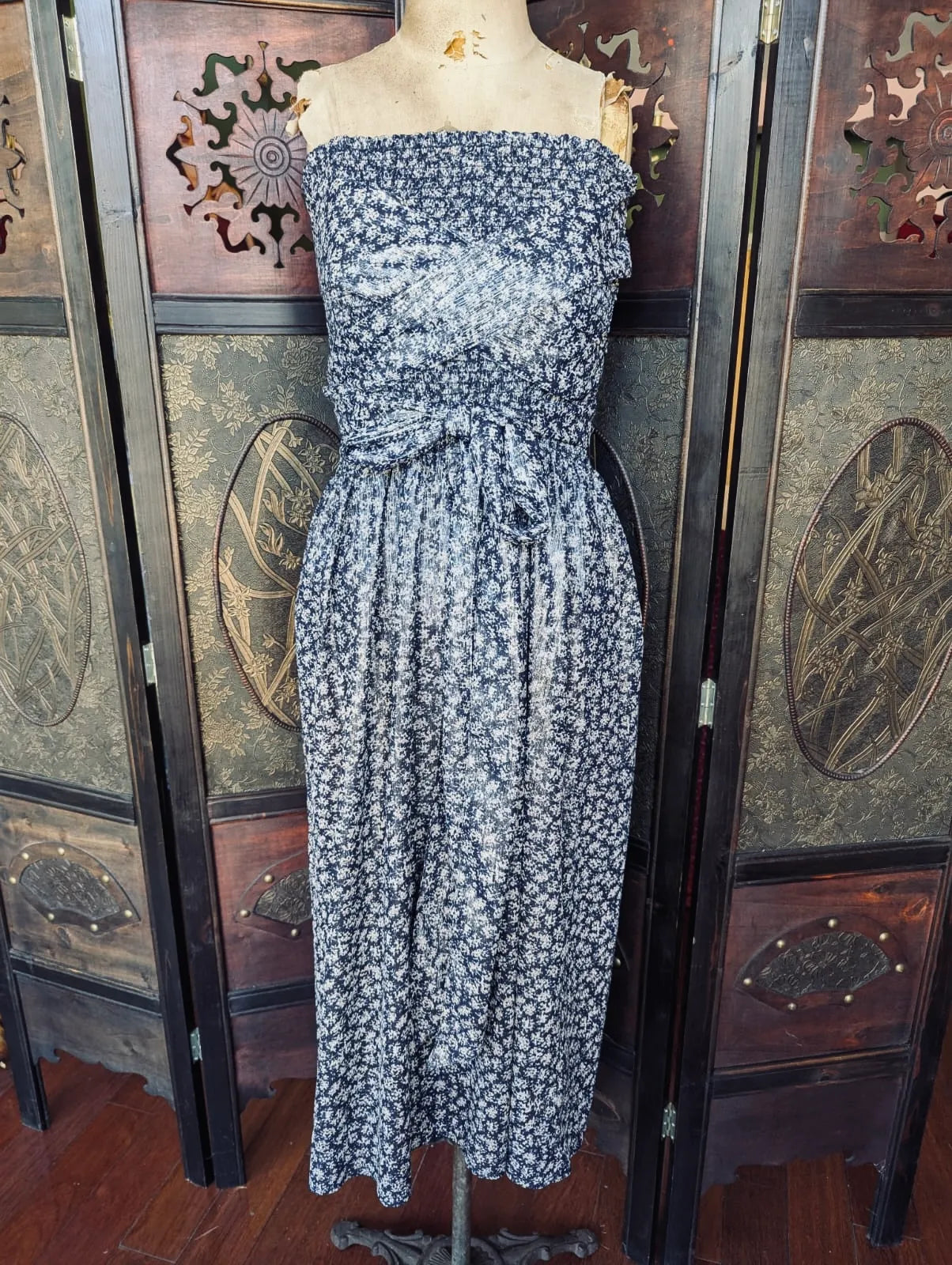 A Broochella Ciao Lucia! strapless midi dress with a small-scale floral print in navy blue and white, displayed on a mannequin in front of a dark wooden folding screen.