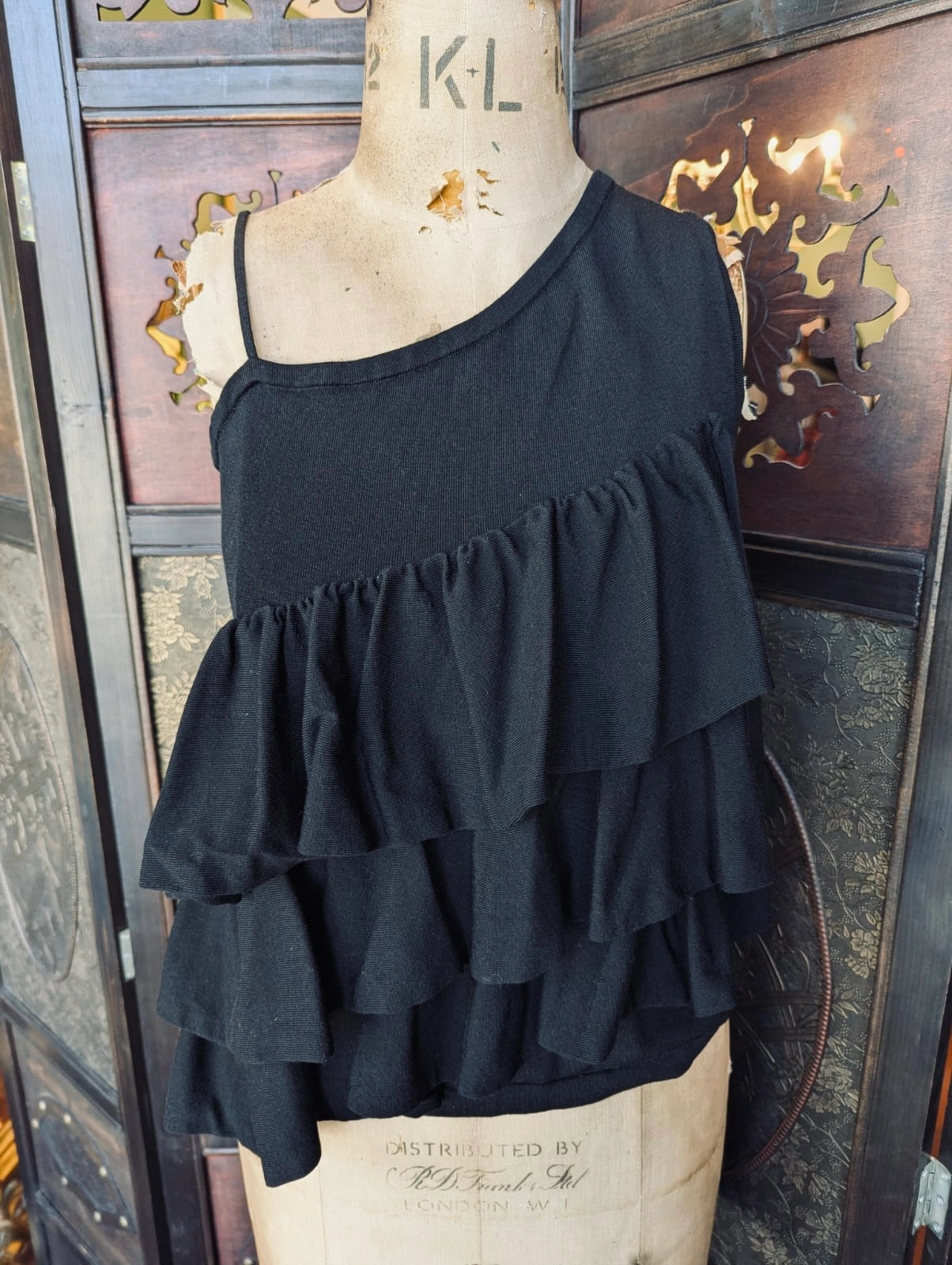 Broochella Co. Black One-Shoulder Tiered knit Ruffle Top – Size Small. It is pictured on a mannequin. The background is an ornate dark wood room divider.