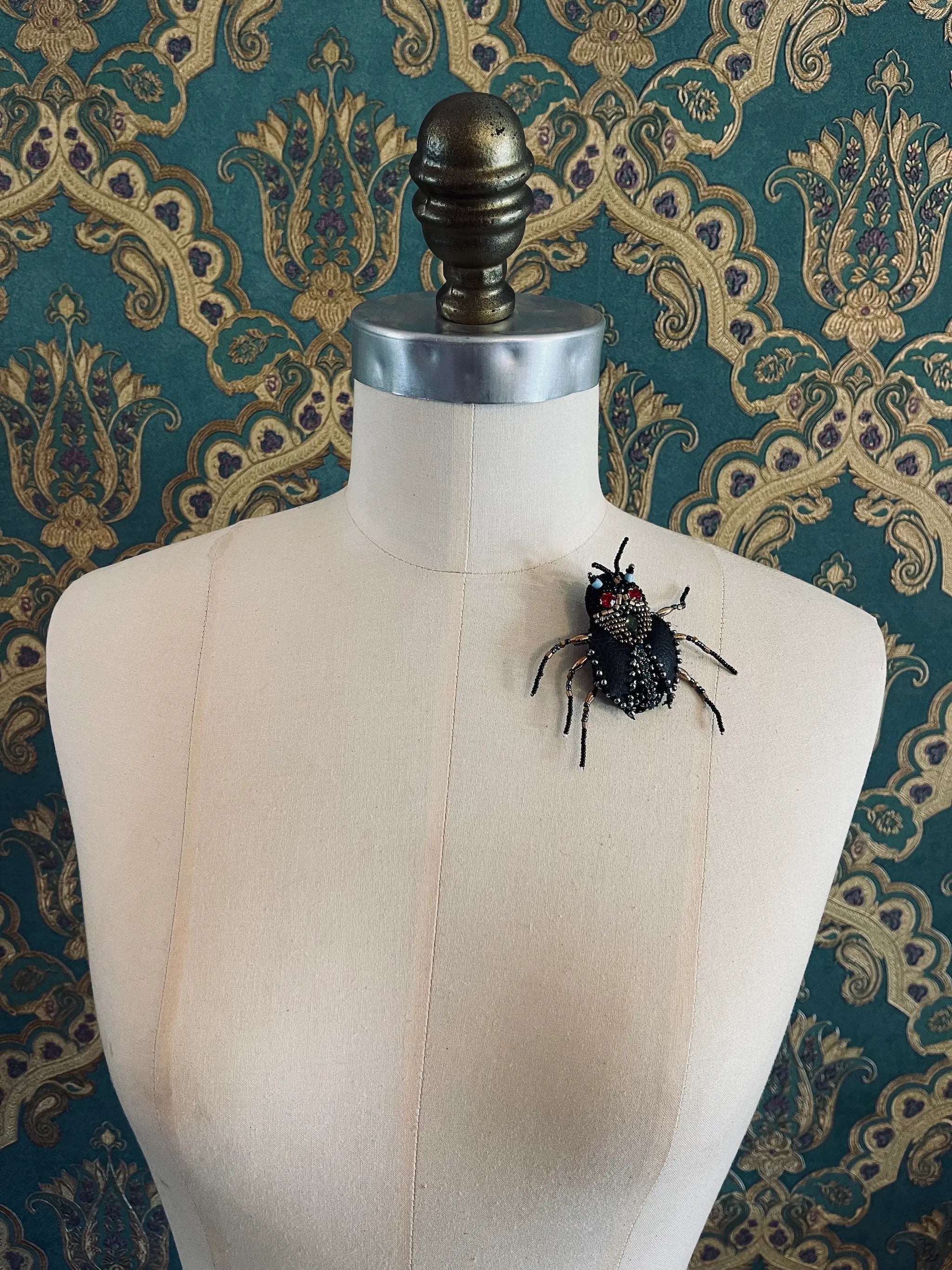 Broochella hand beaded Cockroach brooch in black. Pictured on a mannequin. The background is green and gold damask wallpaper