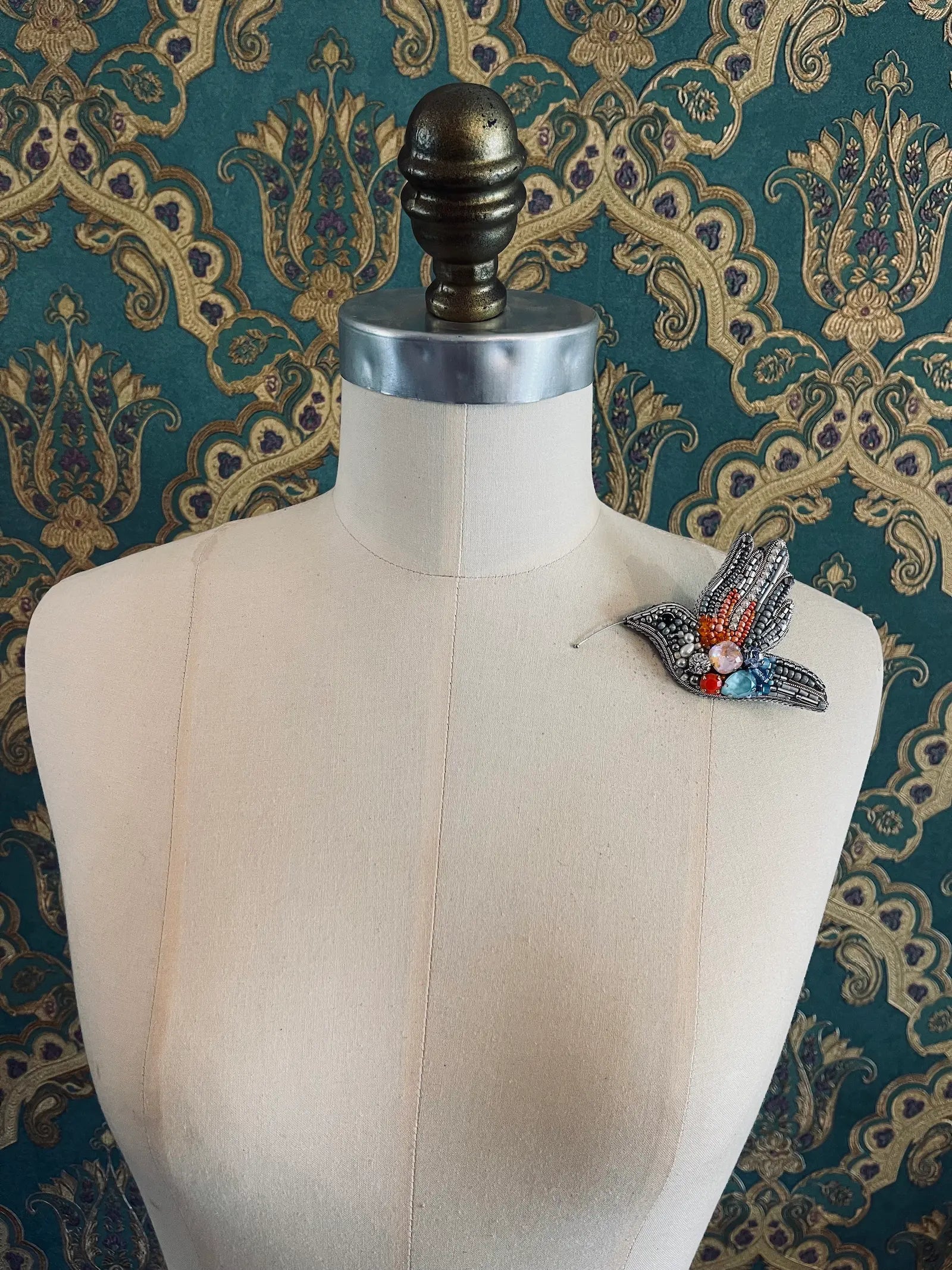 Broochella Columba hand beaded dove bird brooch in blue silver and orange. Pictured on a mannequin. The background is green and gold damask wallpaper.