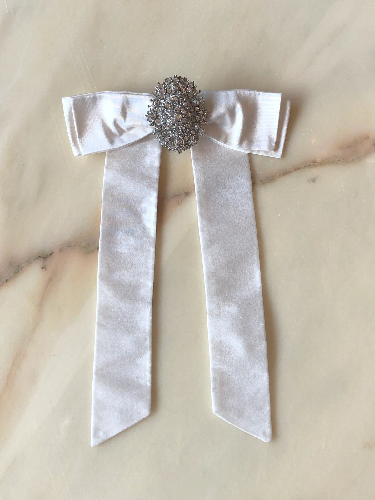 Broochella Contessa Embellished Silk Bow Brooch in ivory, featuring a lustrous silk bow with long tails and an ornate crystal embellishment at the centre, photographed on a pale cream marble background with soft grey and blush veining.