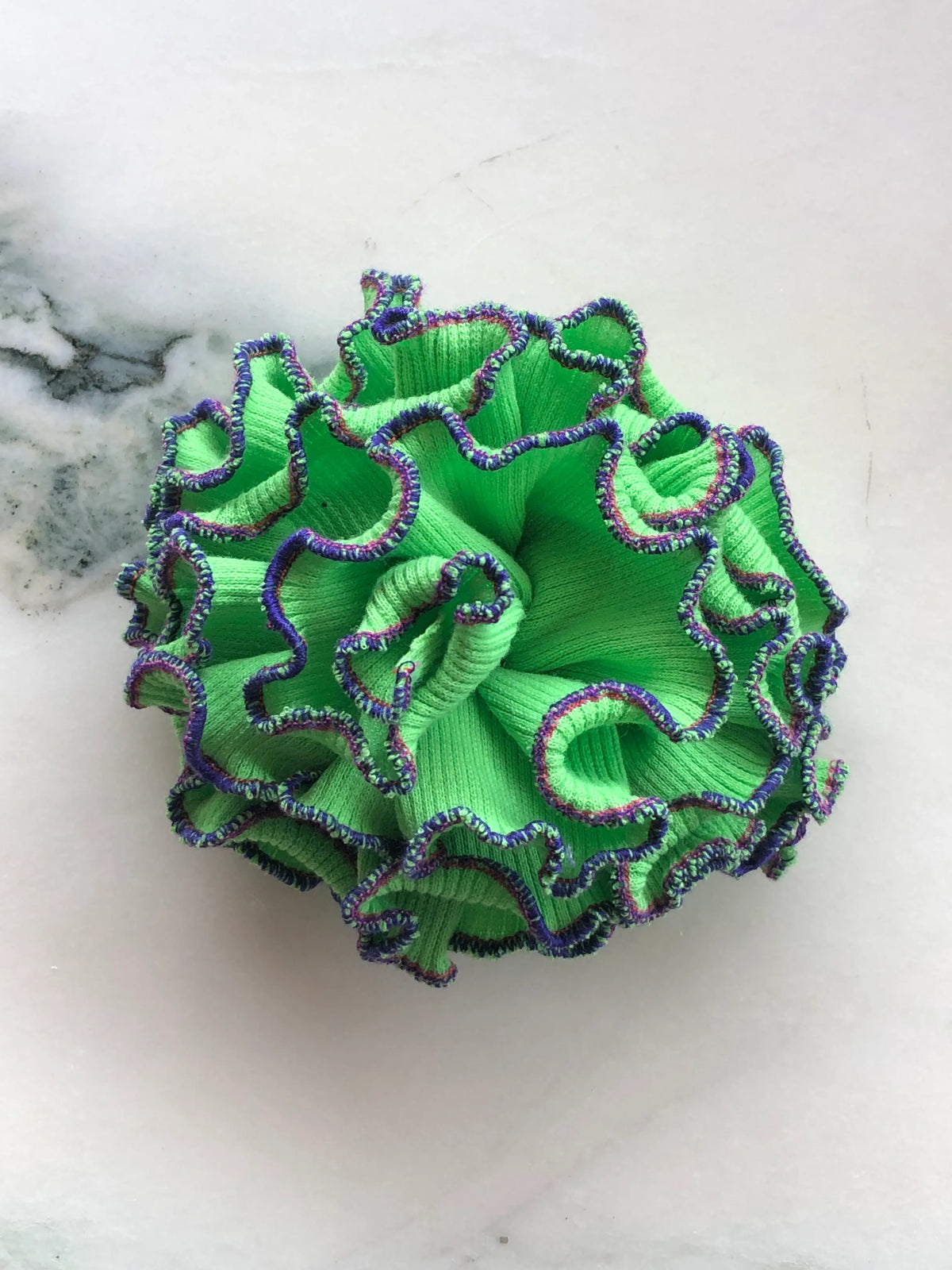 Broochella Corallo Brooch in vivid green with contrasting purple and green ruffled edging, photographed on a white marble background.