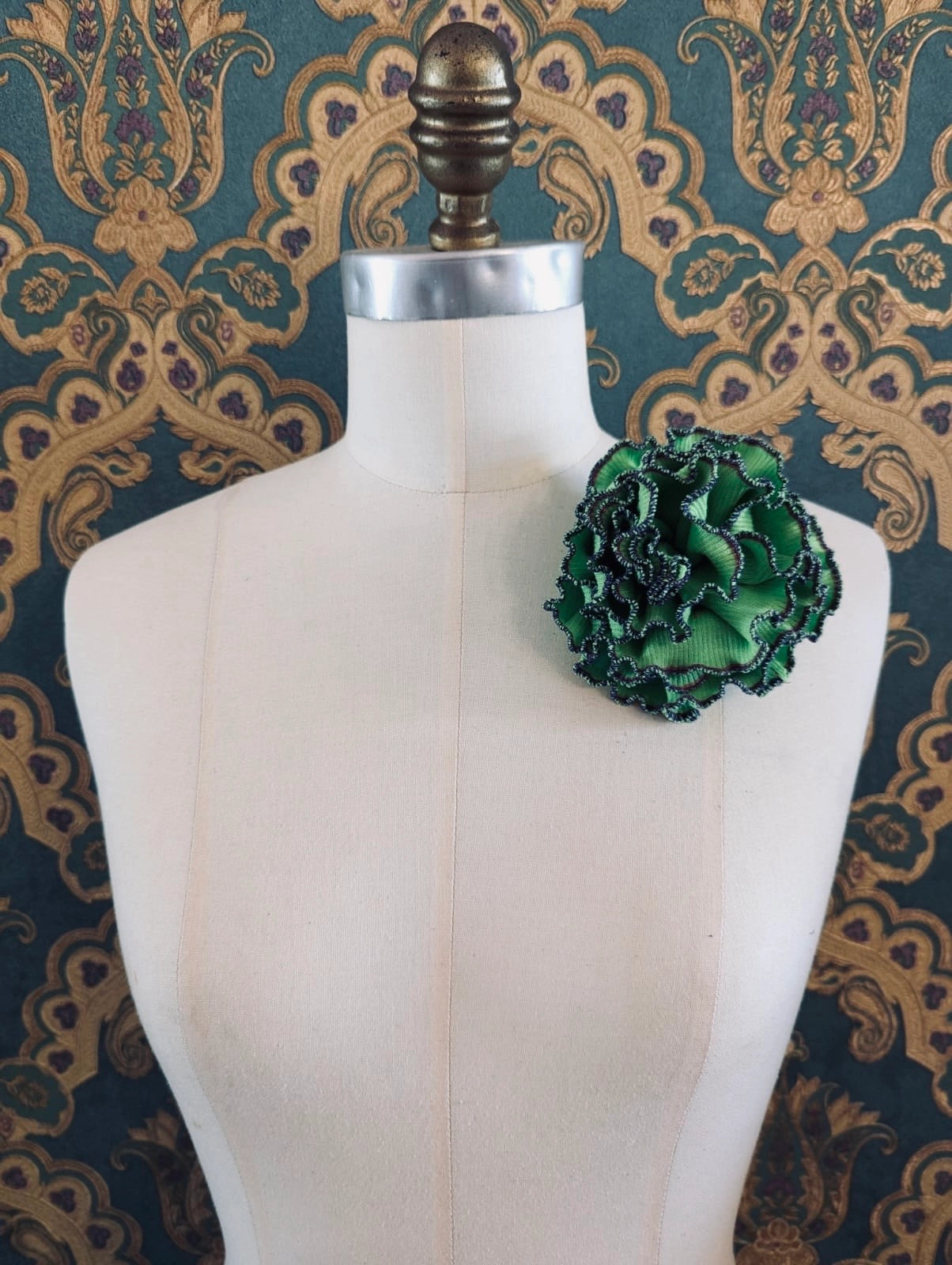 Broochella Corallo handmade fabric flower coral brooch. Made from green stretch fabric, with a blue stitched edge. pinned to a mannequin. The background is green and gold damask wallpaper. 