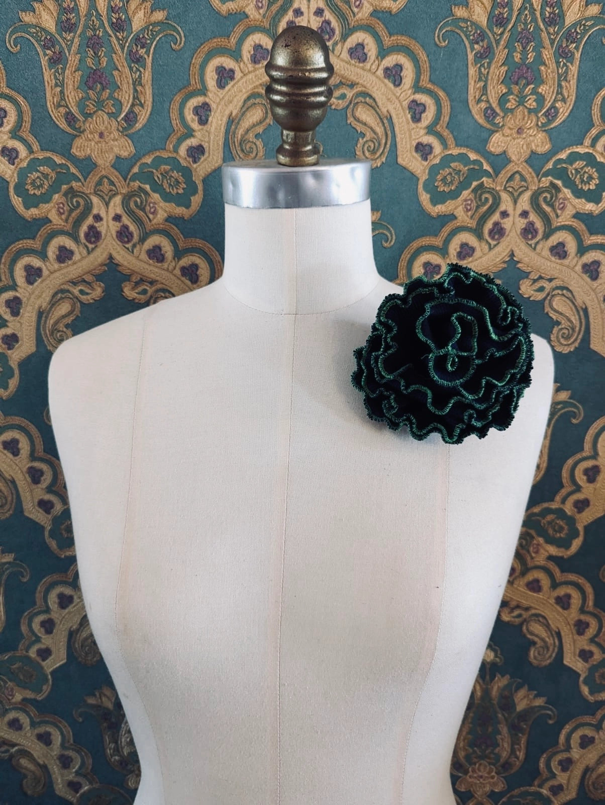 Broochella Corallo handmade fabric flower coral brooch. Made from nay stretch fabric, with a green stitched edge. pinned to a mannequin. The background is green and gold damask wallpaper. 