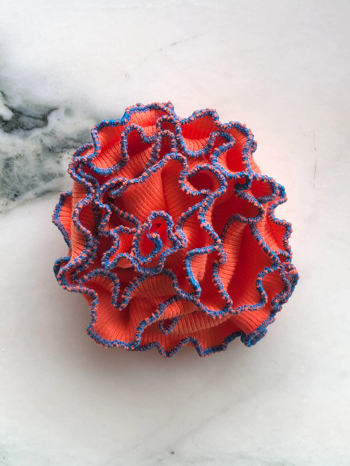 Broochella Corallo Brooch in vibrant orange with contrasting blue and orange ruffled edging, photographed on a white marble background.