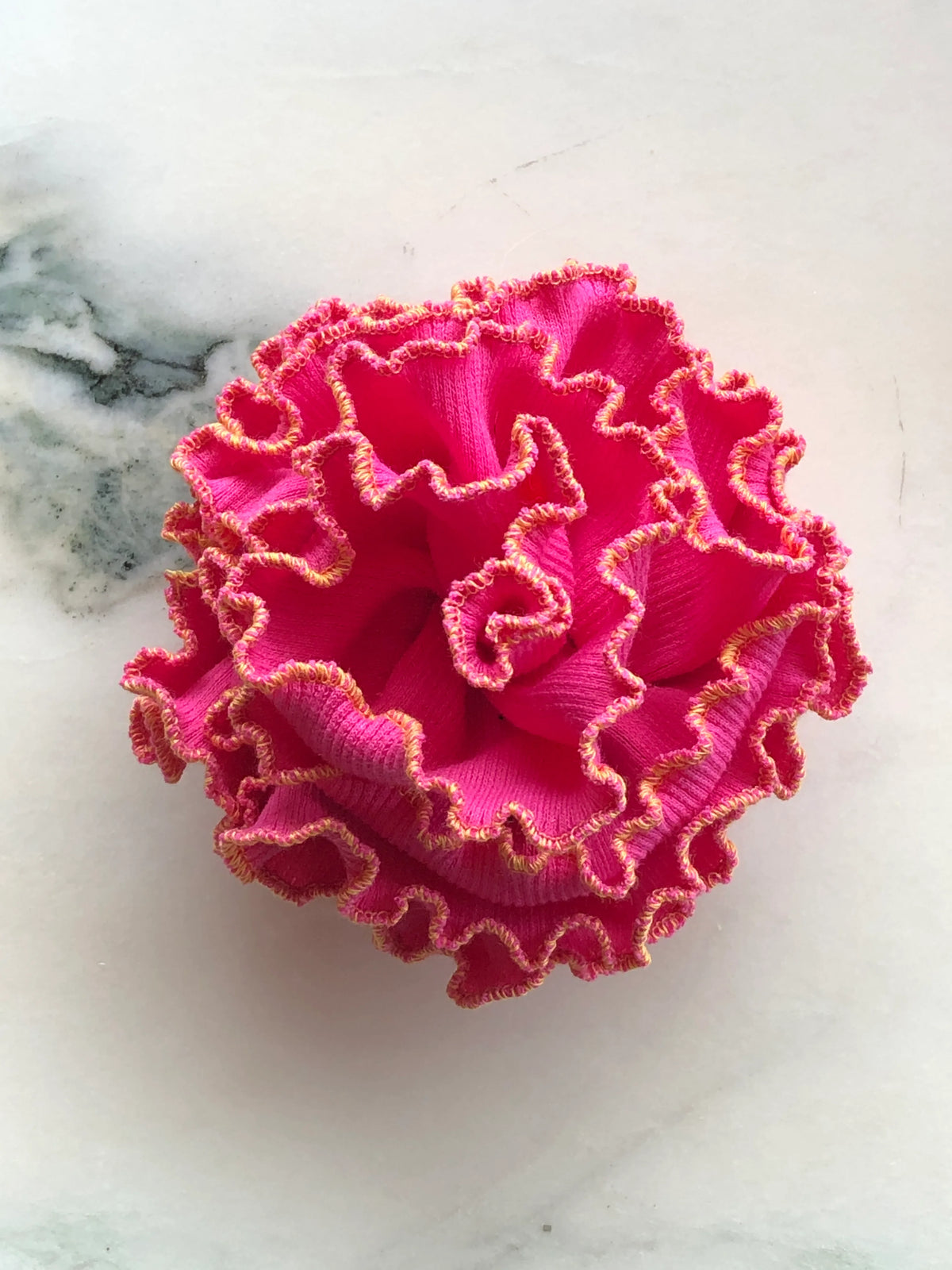 Broochella Corallo Brooch in vibrant pink with contrasting orange-pink ruffled edging, photographed on a white marble background.