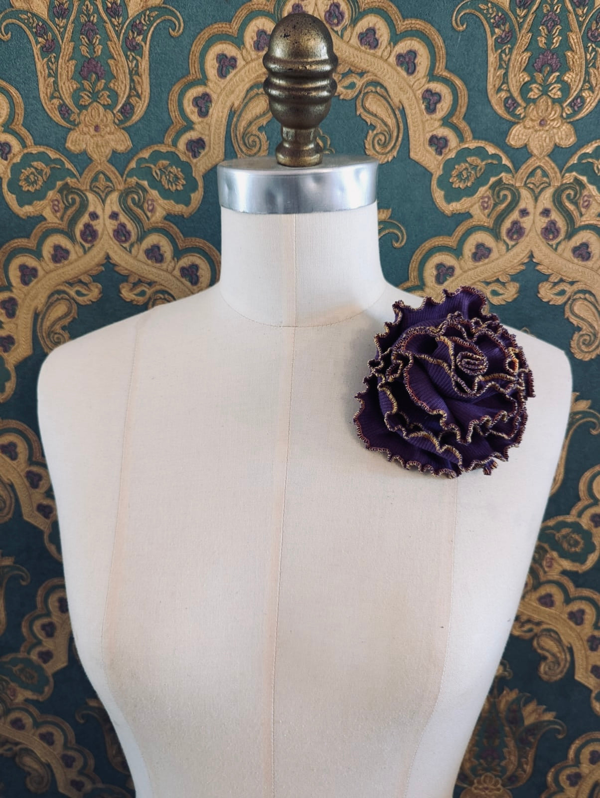 Broochella Corallo handmade fabric flower coral brooch. Made from purple stretch fabric, with a yellow and pink stitched edge. pinned to a mannequin. The background is green and gold damask wallpaper. 