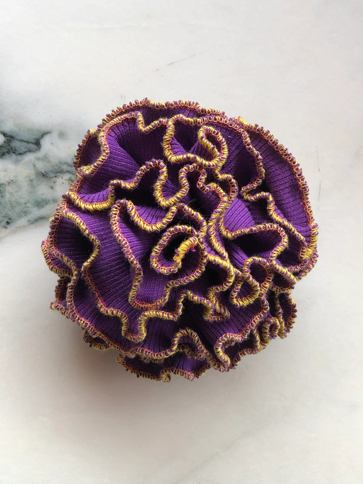 Broochella Corallo Brooch in rich purple with contrasting yellow and purple ruffled edging, photographed on a white marble background.