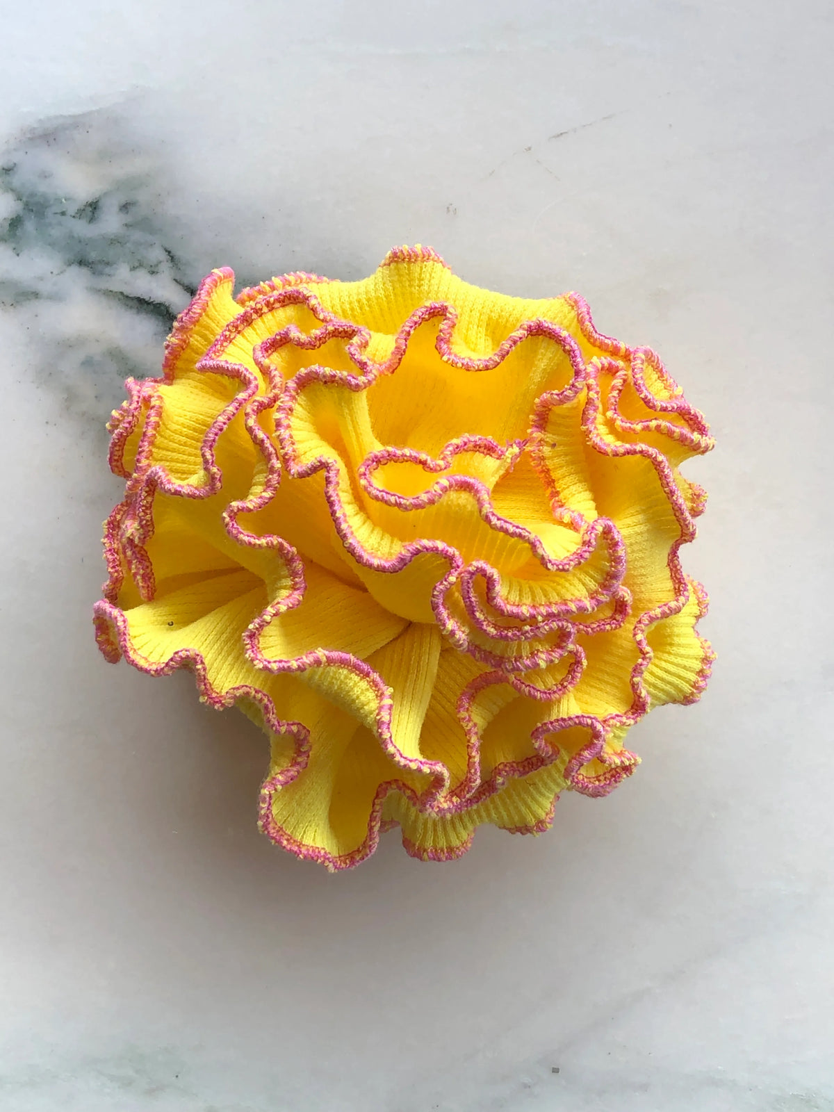 Broochella Corallo Brooch in bright yellow with contrasting pink ruffled edging, photographed on a white marble background.