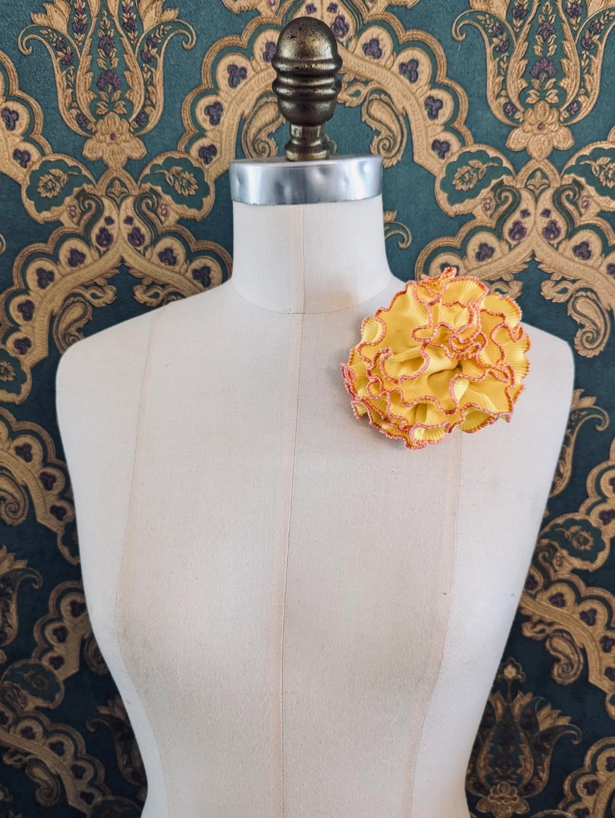 Broochella Corallo handmade fabric flower coral brooch. Made from yellow stretch fabric, with a pink stitched edge. pinned to a mannequin. The background is green and gold damask wallpaper. 