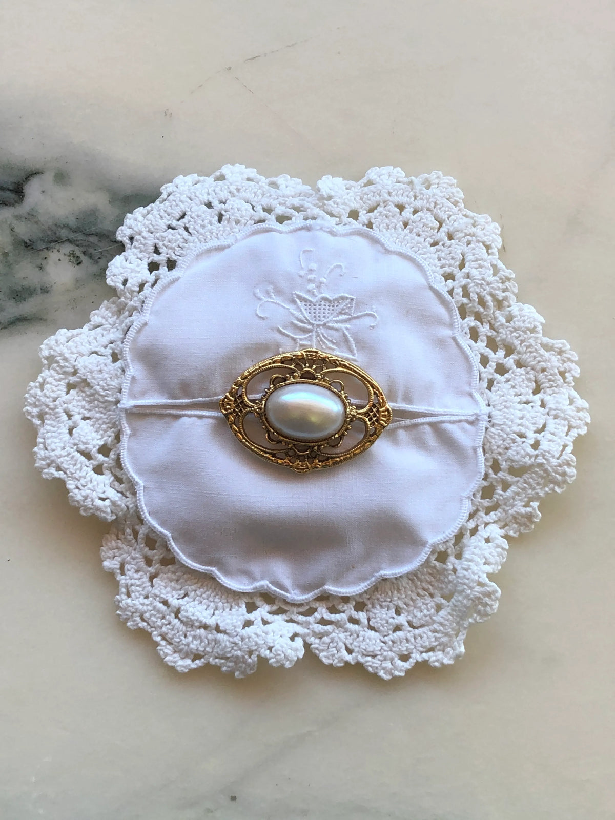 Broochella Corredi brooch pin handcrafted from a white vintage crochet doily and embroidered scalloped linen centre, finished with an ornate gold-tone vintage brooch featuring a large luminous pearl-style centre, photographed on a white marble background with soft grey-green veining.