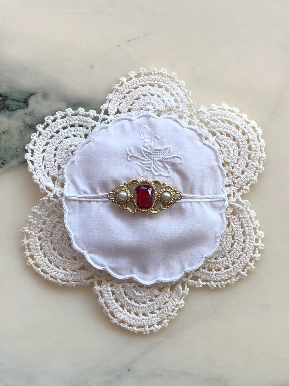 Broochella Corredi brooch pin handcrafted from white vintage crochet doilies and an embroidered scalloped linen centre, finished with an ornate gold-tone vintage brooch featuring a rectangular ruby-red stone and two pearl accents, photographed on a white marble background with soft grey-green veining.