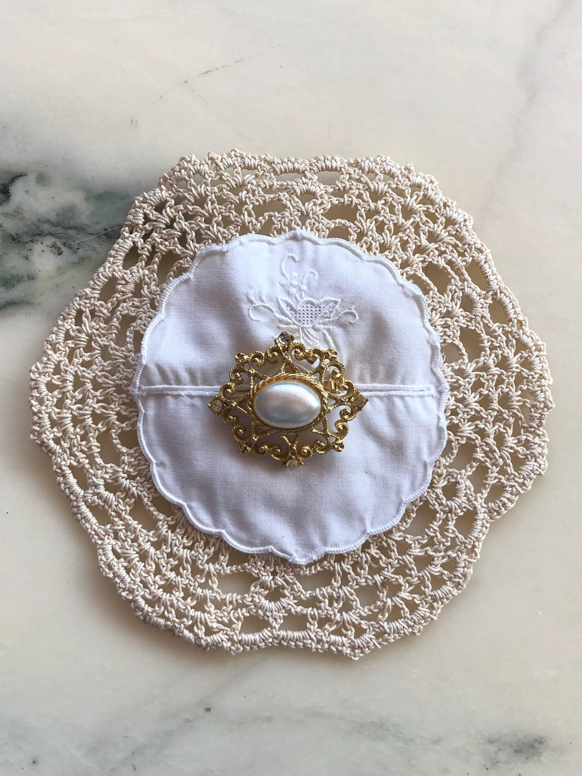 Broochella Corredi brooch pin handcrafted from an ivory crochet vintage doily layered with a white embroidered scalloped doily, centred with an ornate gold-tone vintage brooch featuring a large oval pearl, photographed on a white marble background with soft grey-green veining.