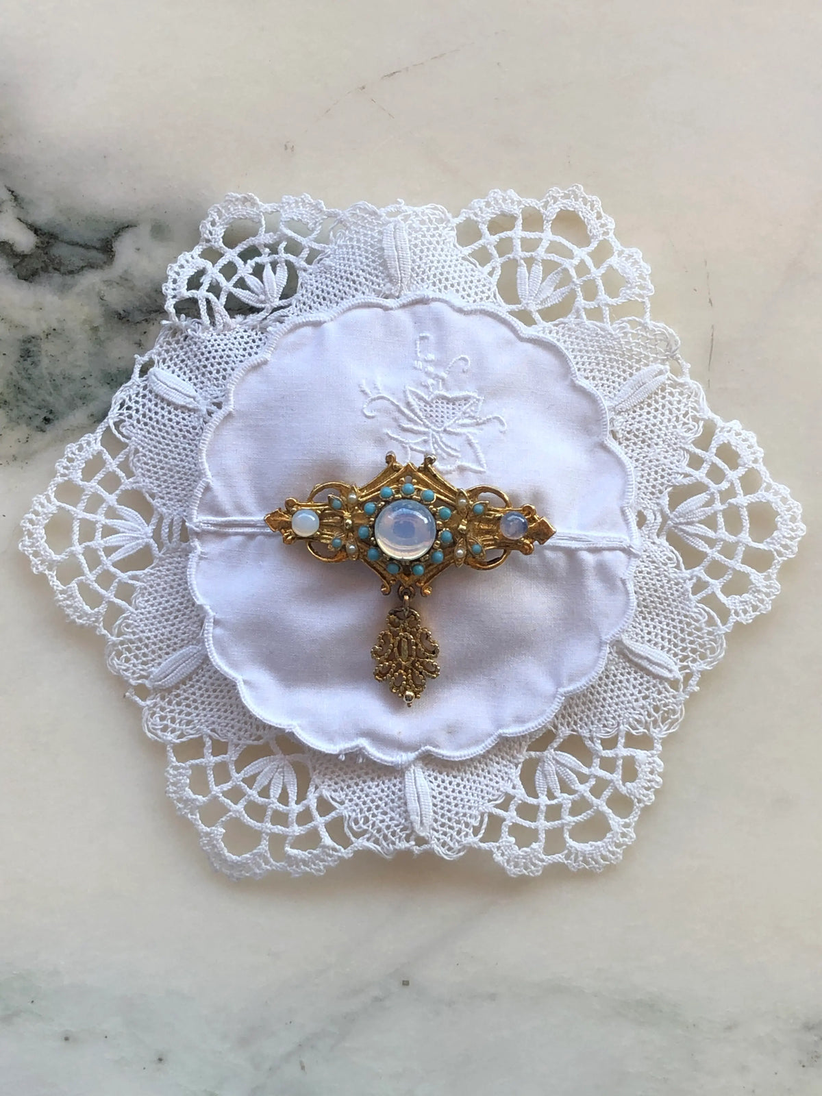 Broochella Corredi brooch pin handcrafted from a white vintage embroidered doily, finished with an ornate gold-tone vintage brooch featuring opalescent and turquoise stones, photographed on a white marble background with soft grey-green veining.