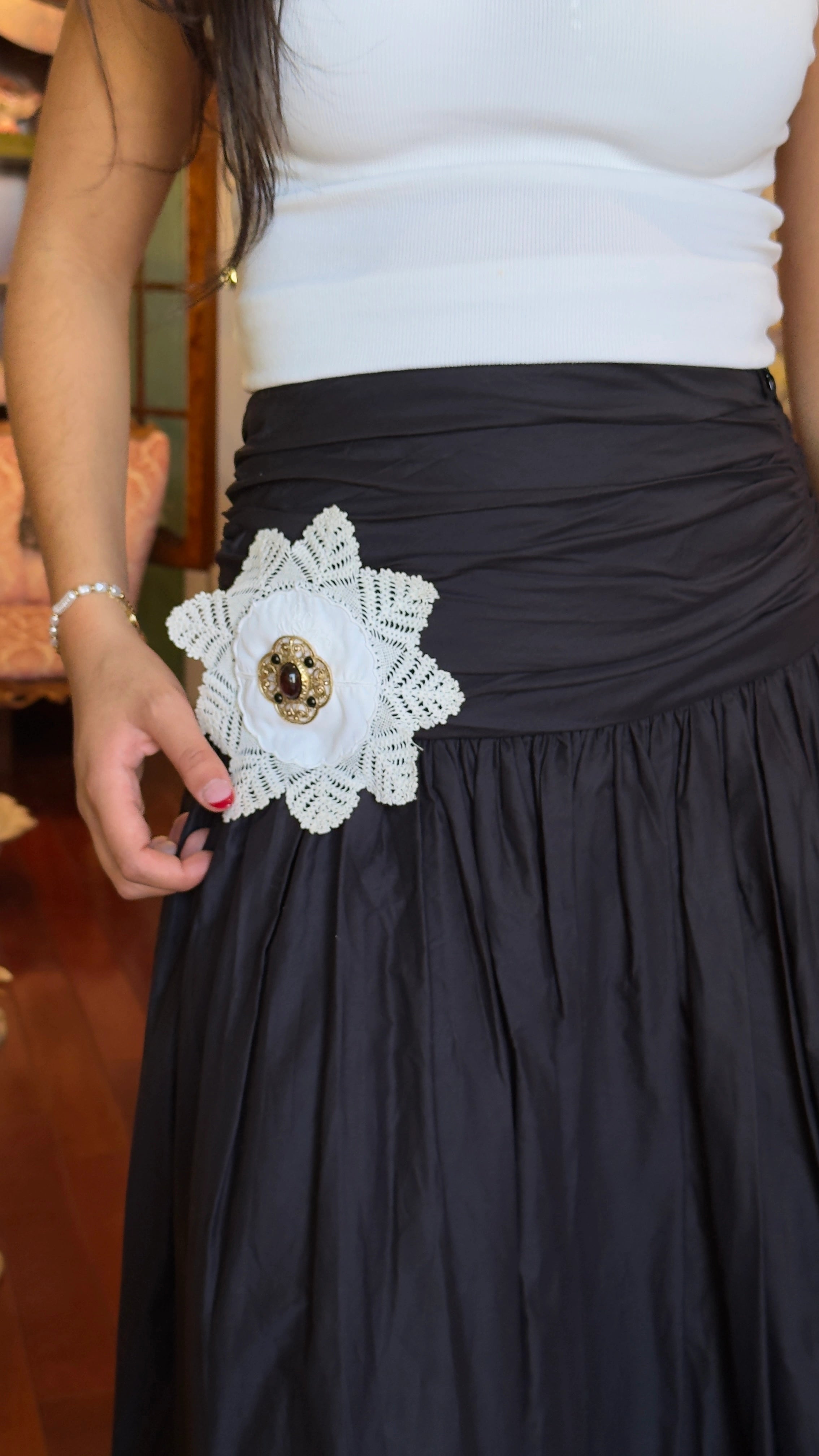 A close-up of a person wearing a black gathered skirt with a large white Broochella Corredi 4/11 vintage doily brooch featuring a gold-toned vintage brooch embellishment, paired with a white top.