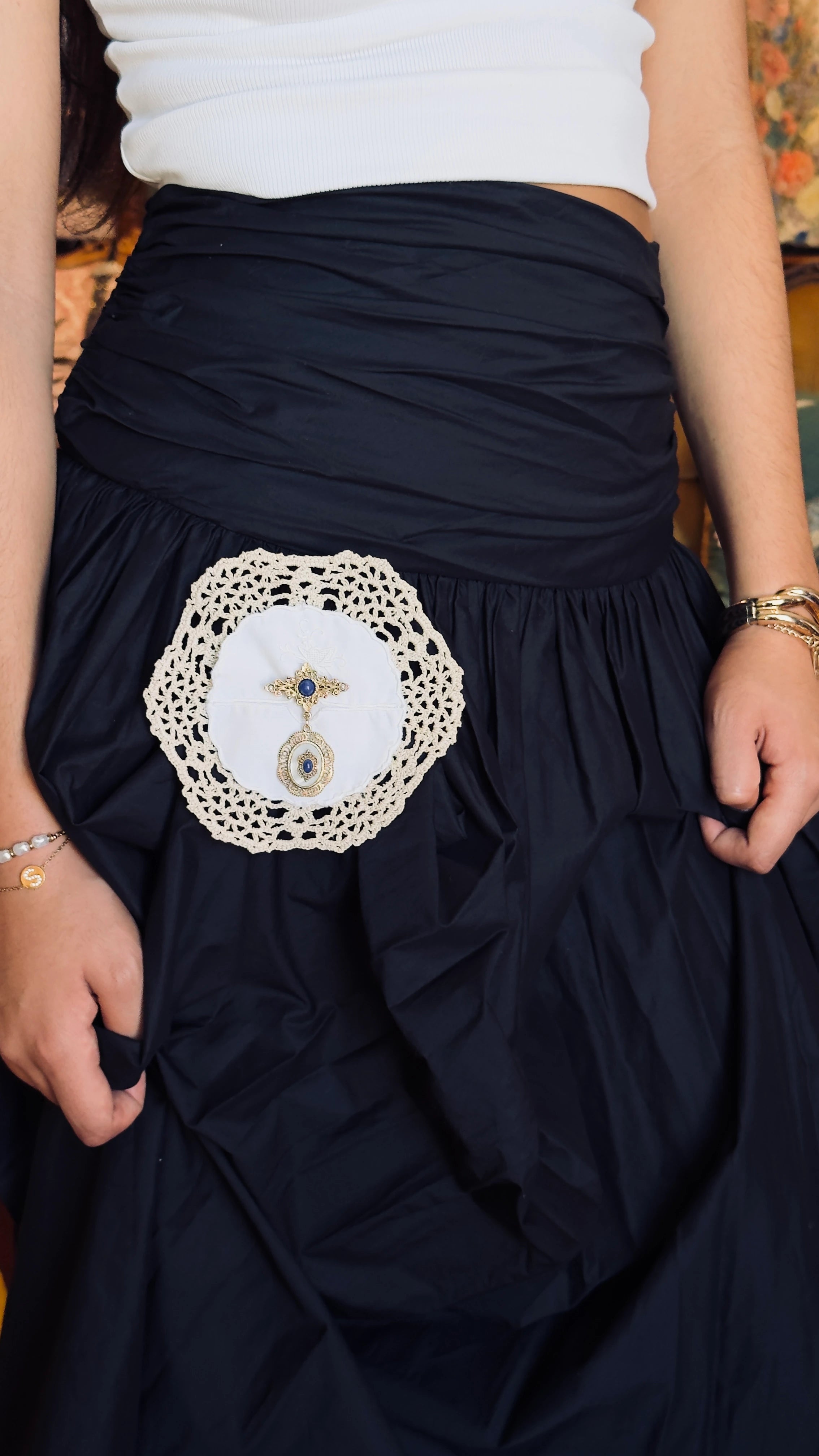 A close-up of a person wearing a black skirt with a decorative Broochella Corredi  white line, and lace doily brooch. It features a central teardrop-shaped brooch embellishment.