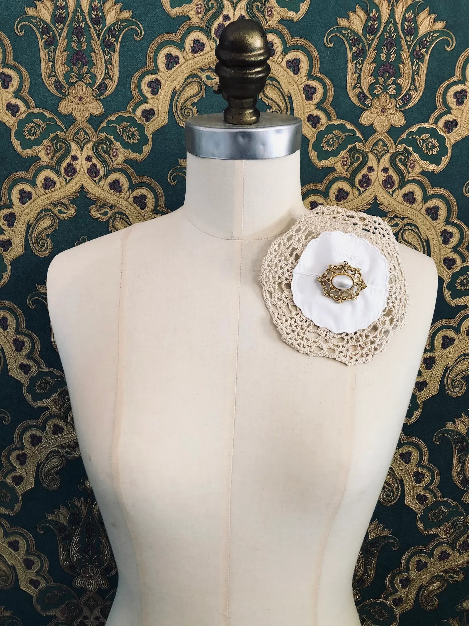 Broochella Corredi 11/11 handmade vintage lace, linen and doily brooch. The centre embellishment is a vintage gold tone metal brooch, with a blue stone centre. It is pinned to a mannequin. The background is green and gold damask wallpaper. 