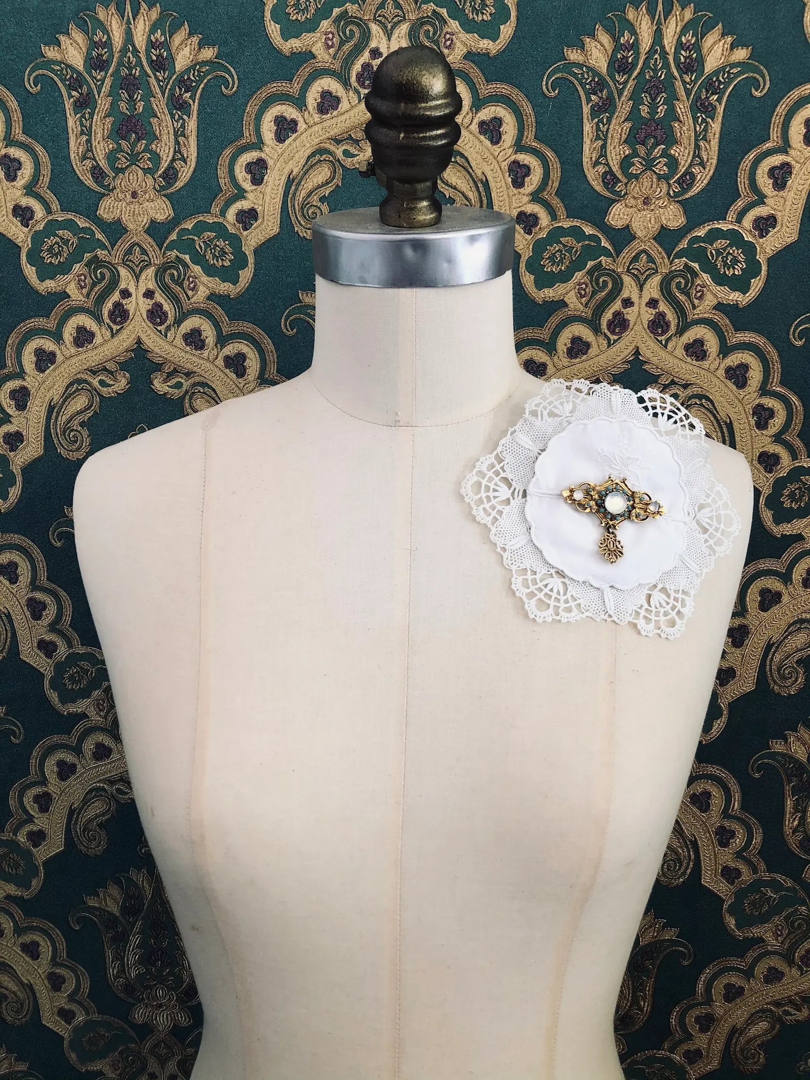 Broochella Corredi 2/11 vintage white doily and lace brooch with a vintage brooch embellishment. It is pinned to a mannequin. The background is green and gold damask wallpaper.