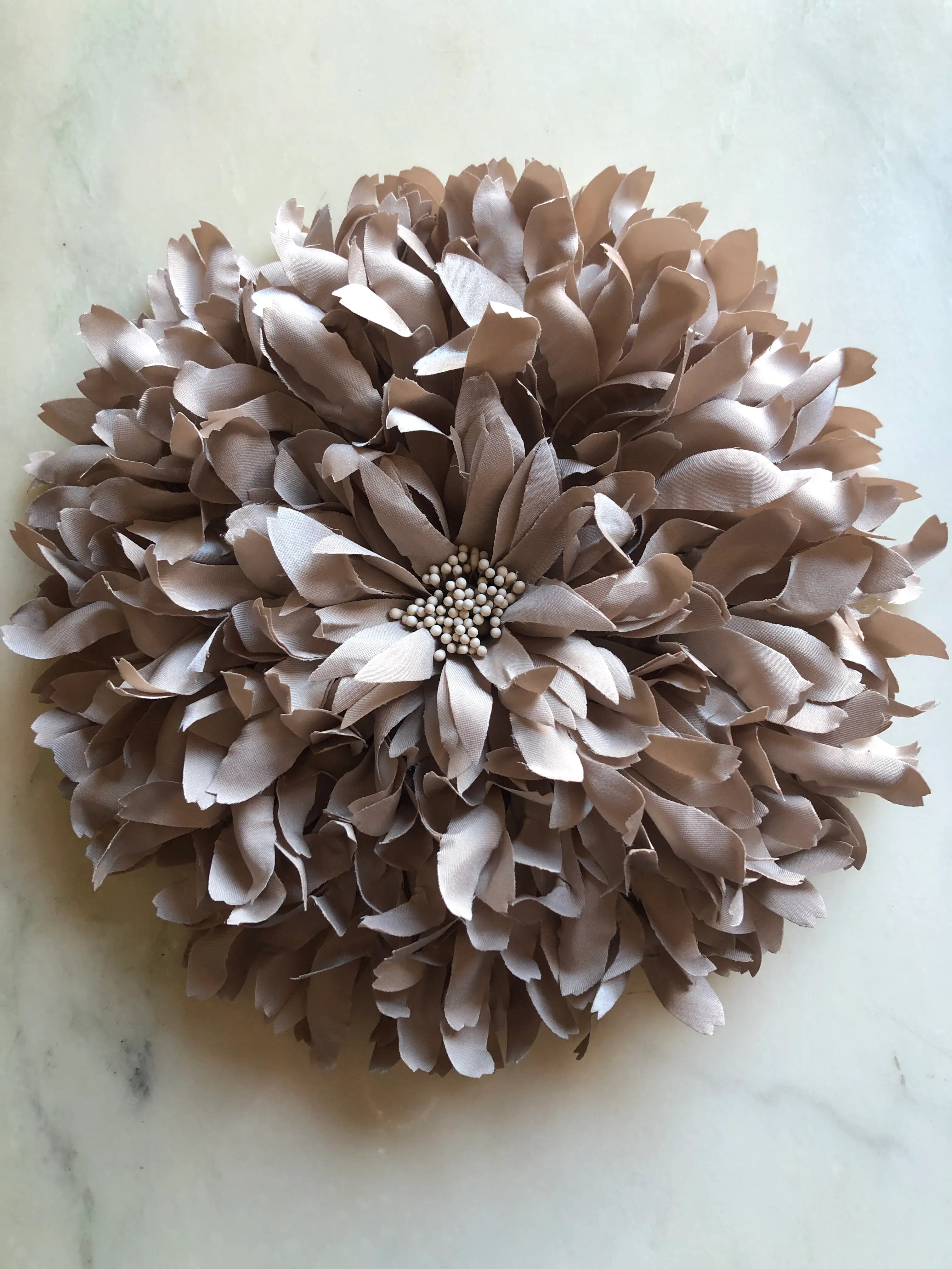 A  Broochella Crisantemo large a flower brooch with layered taupe-brown and beige petals, arranged in a round pom-pom style, with a small cluster of pearl-like beads at the center, displayed on a light marble surface.