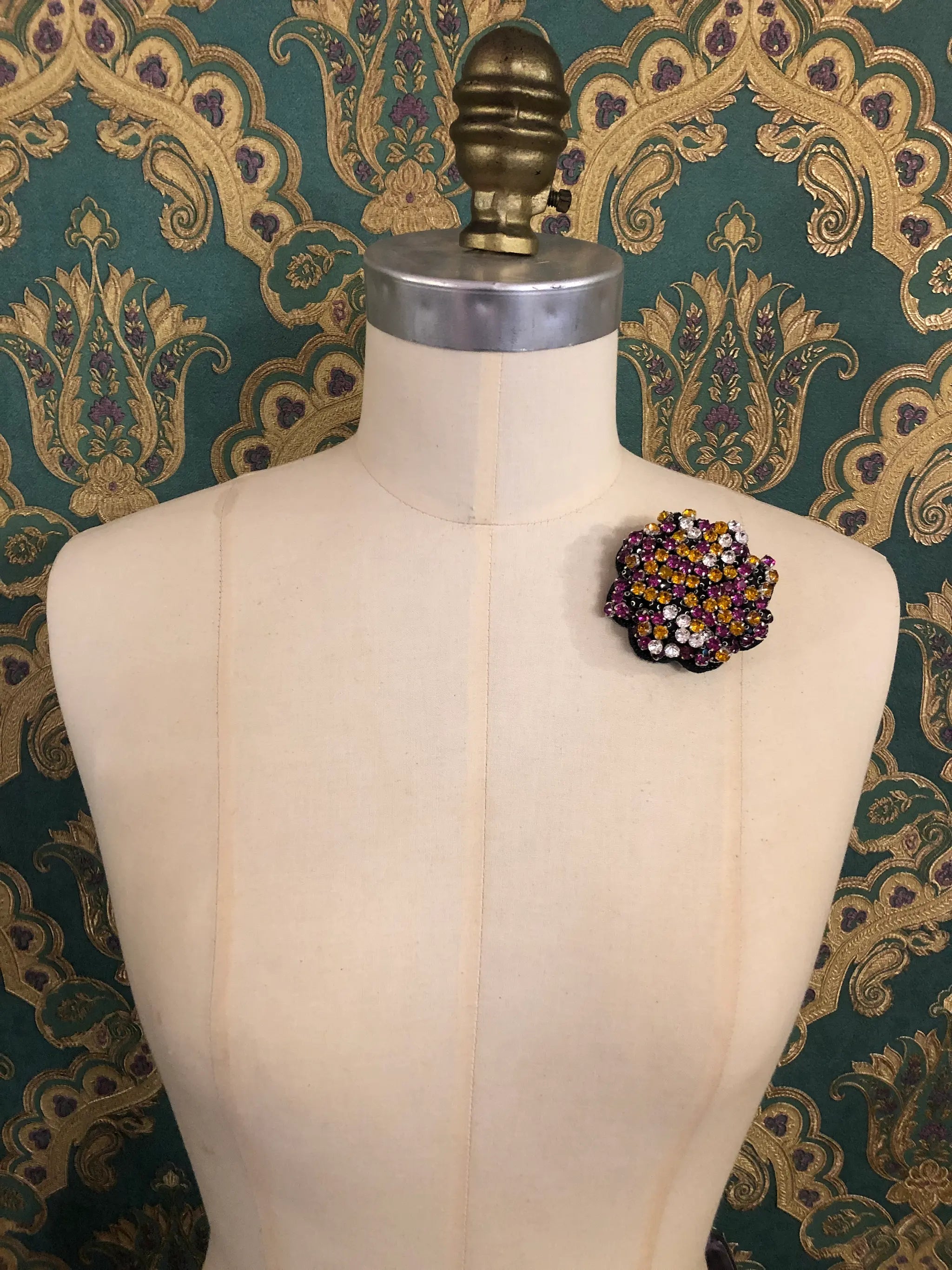 A Broochella Crystal Blooms small floral brooch with magenta, orange, and white rhinestones, displayed on a cream-colored dress form against a dark green and gold patterned wallpaper background.
