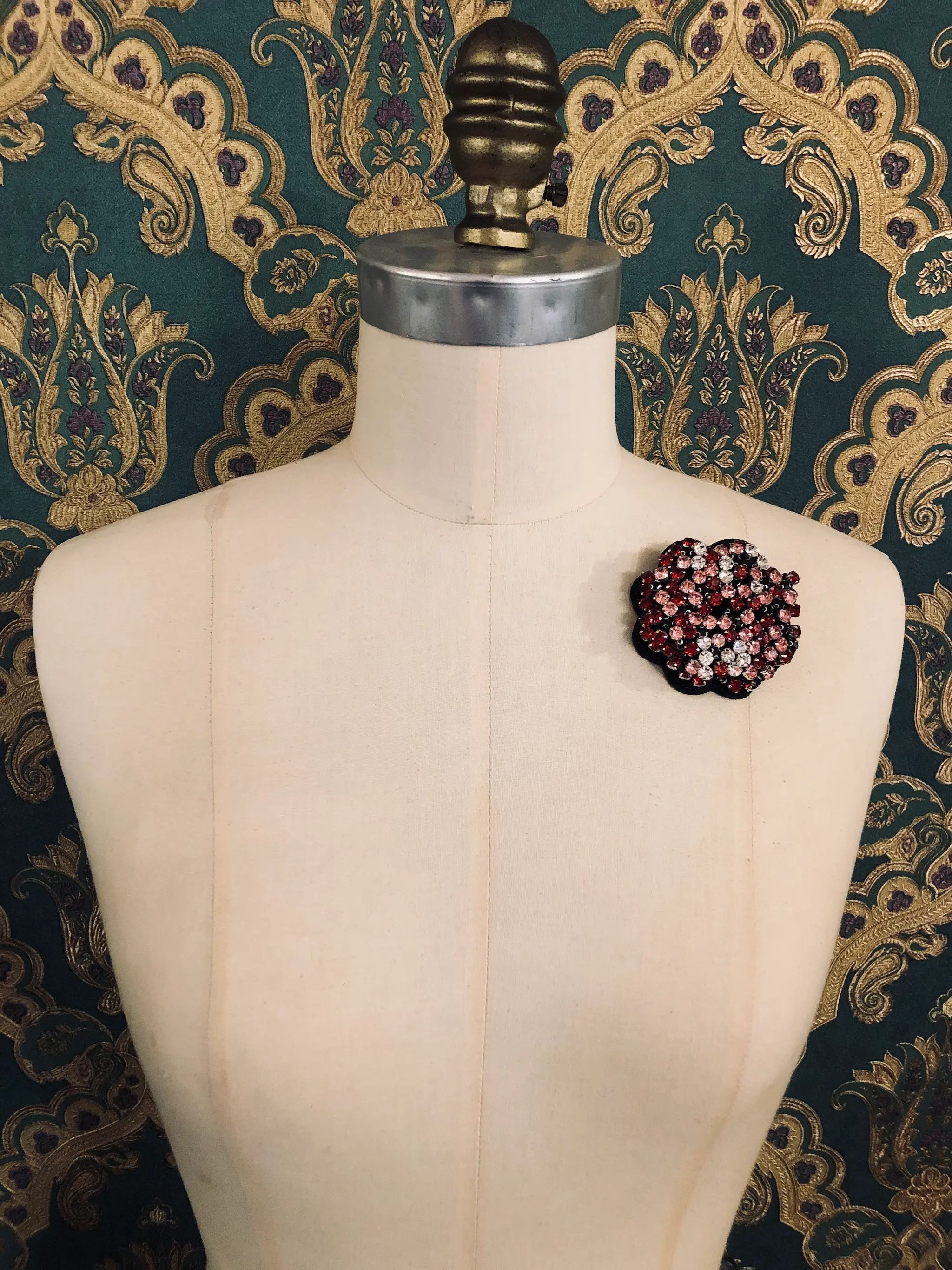 A Broochella Crystal Blooms small floral brooch with dark red, pink, and white rhinestones, displayed on a cream-coloured dress form against a dark green and gold patterned wallpaper background.