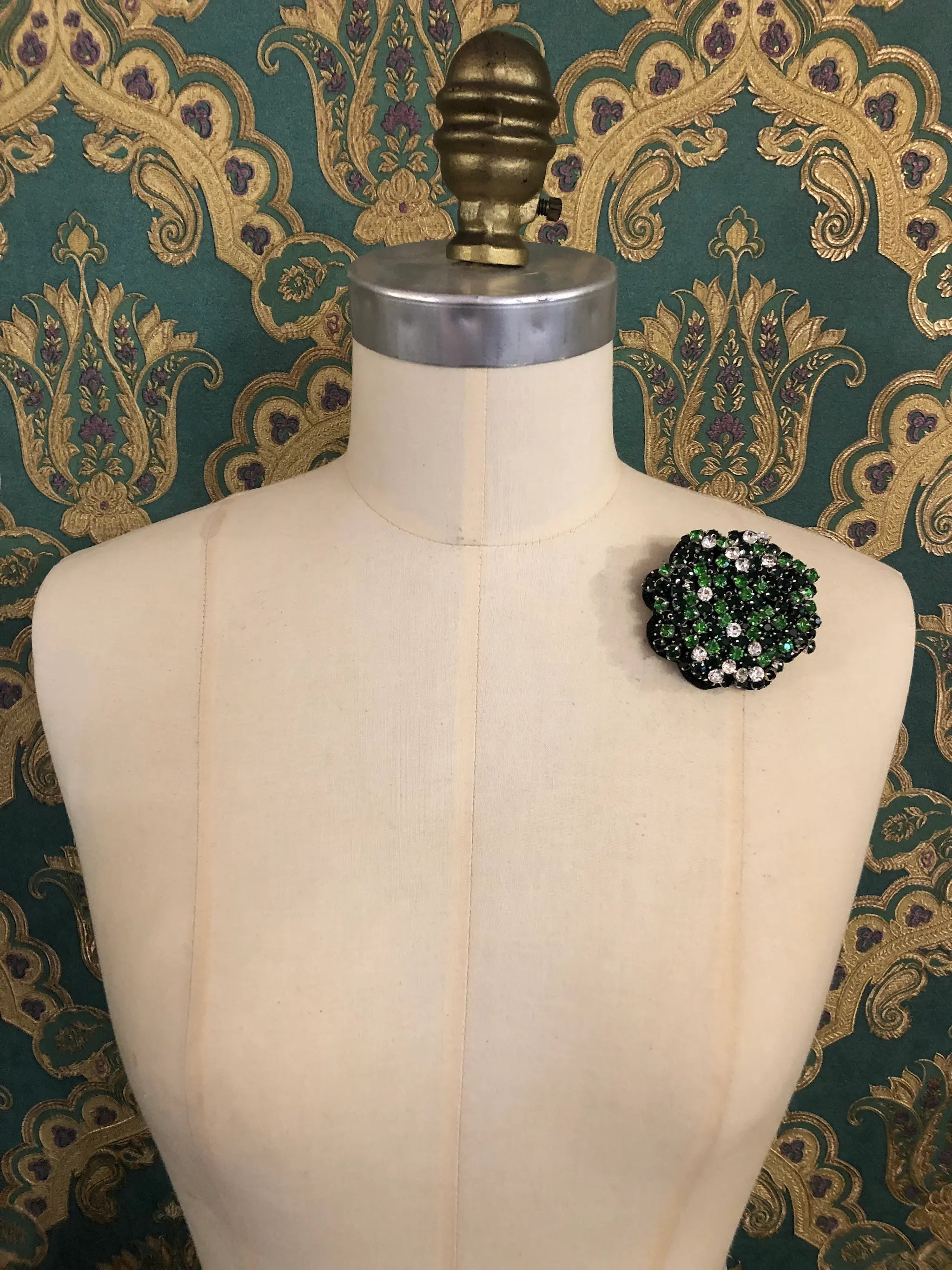 A Broochella Crystal Blooms small floral brooch with dark green, lime, and white rhinestones, displayed on a cream-colored dress form against a dark green and gold patterned wallpaper background.