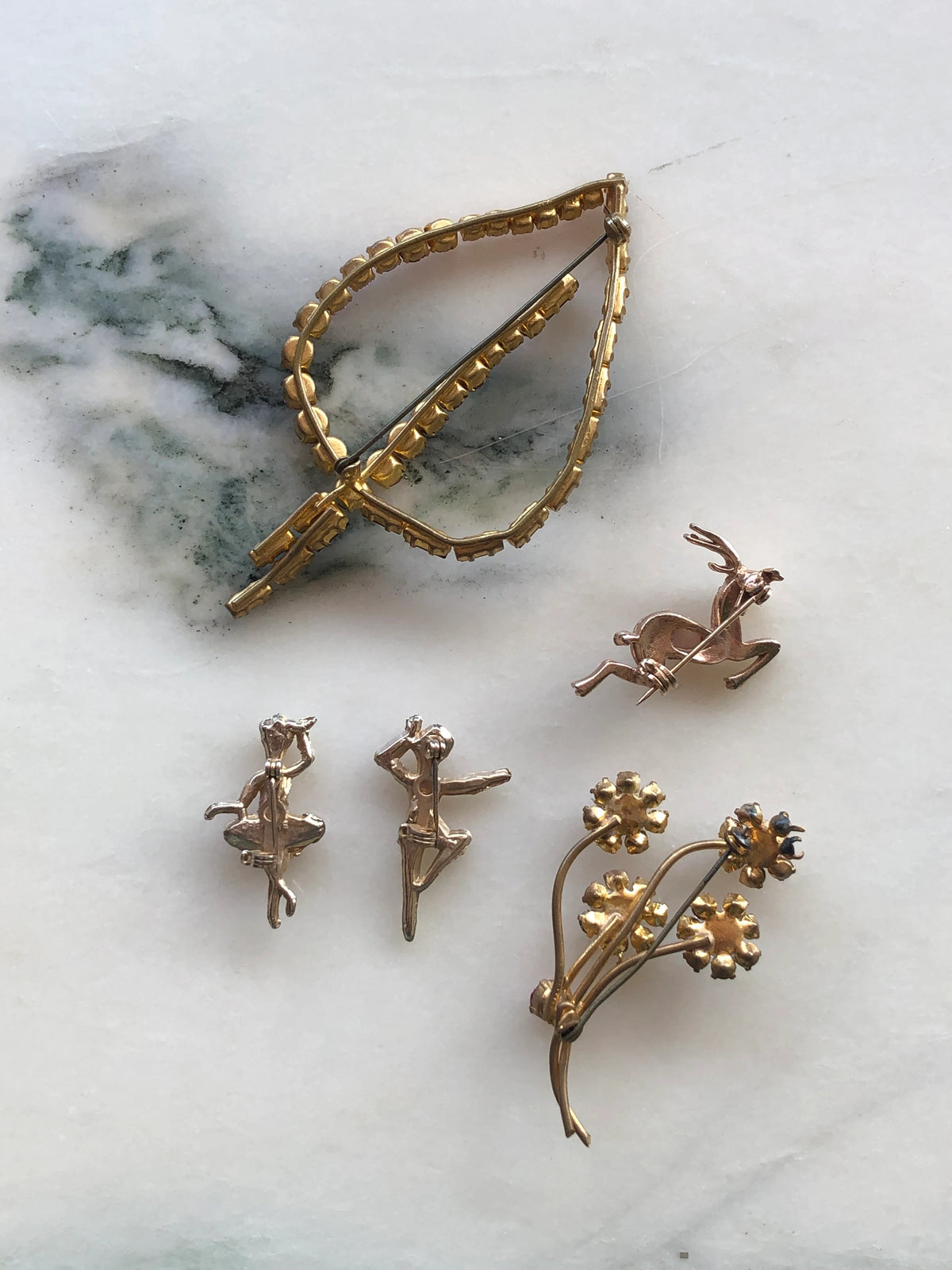 The back view of Broochella Da Vinci Quint Vintage Brooch Set featuring five delicate pink and purple crystal designs including an initial, ballerinas, leaping deer and floral spray, photographed on a white marble background.