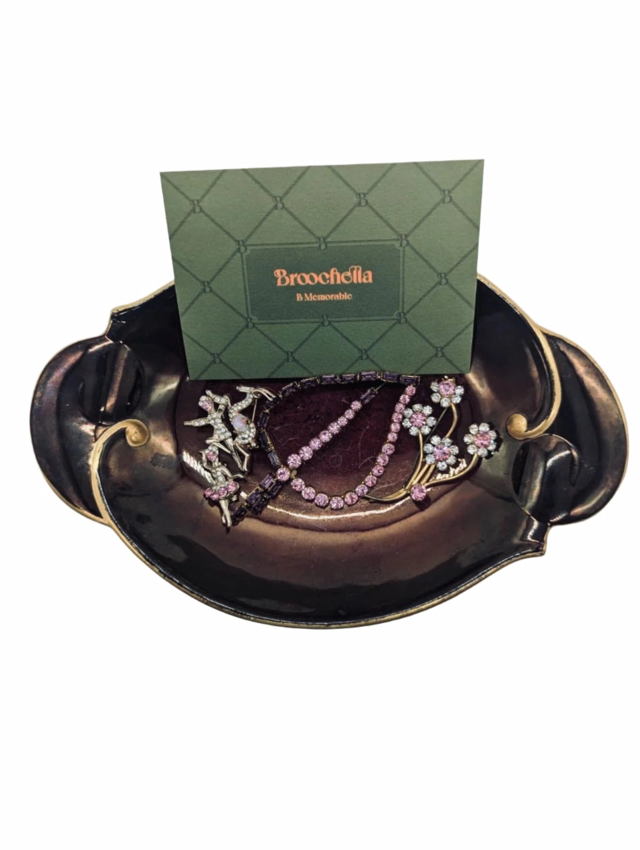 Broochella da vinci five piece vintage brooch set in gold tone metal, with pink crystals. The set includes flowers, dancing figures, a leaf and a deer. They are placed on a wembley ware burgundy and gold tray. There is a broochella business card behind the brooches. The background is white. 