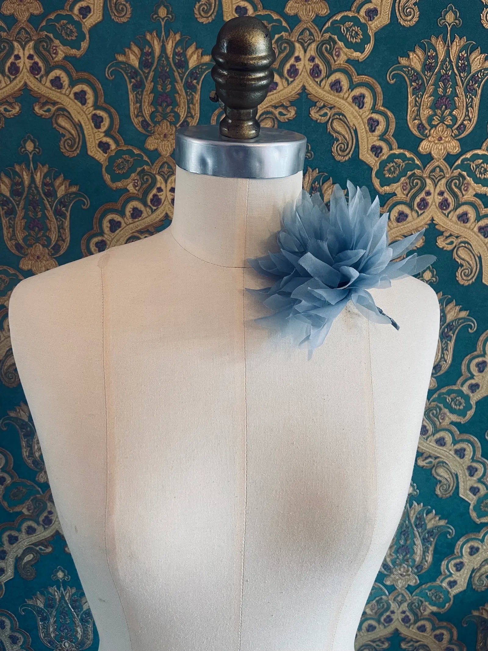 Broochella Dahlia handmade fabric flower brooch in blue. Styled on a mannequin. The background is green and gold damask wallpaper