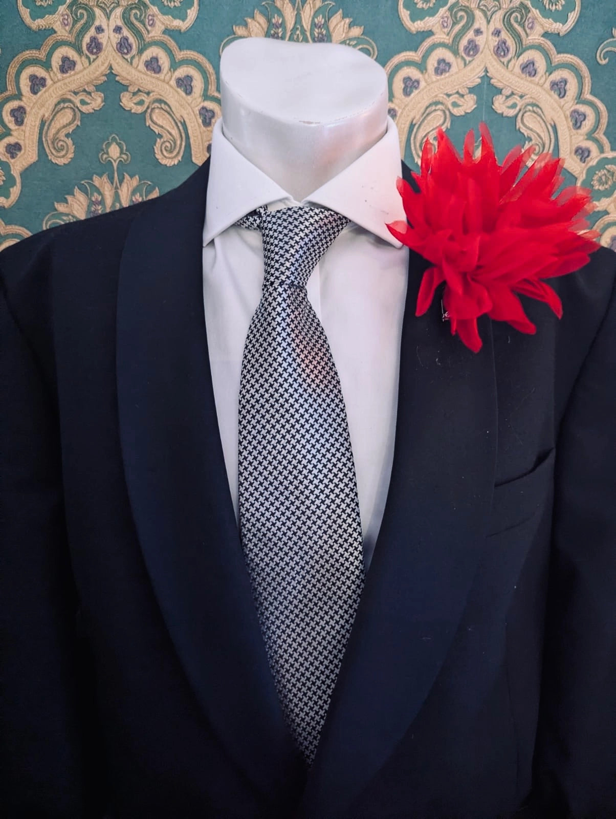 A black suit jacket displayed on a white mannequin torso, styled with a white dress shirt, a patterned grey and white tie, and a red Broochella dahlia flower brooch against a teal and gold damask wallpaper background.