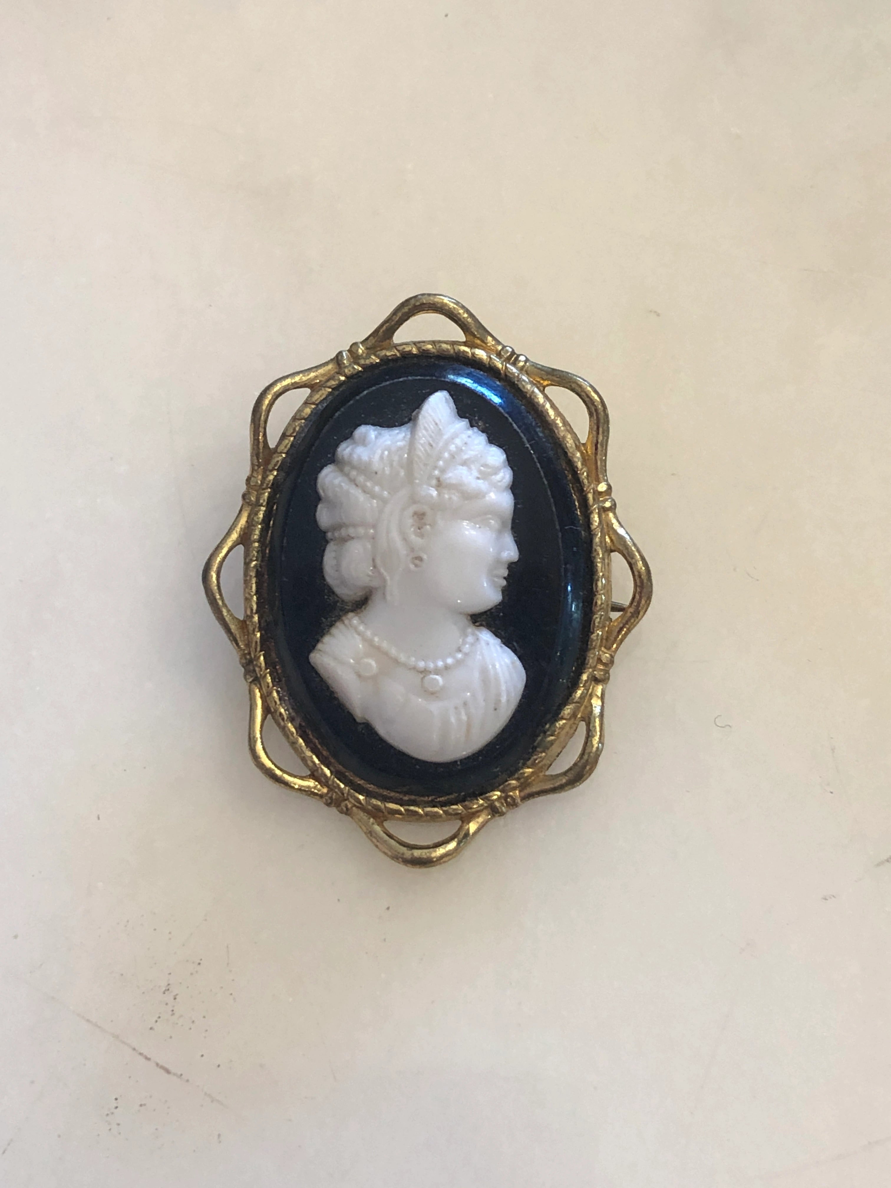 A Broochella Dea 4cm x 3cm vintage black and white cameo brooch featuring a carved white relief classical portrait of a noblewoman in profile with elaborate feathered and pearl upswept hair and a pearl necklace, set on a deep black oval ground within a gold-tone openwork filigree frame. The background is white marble