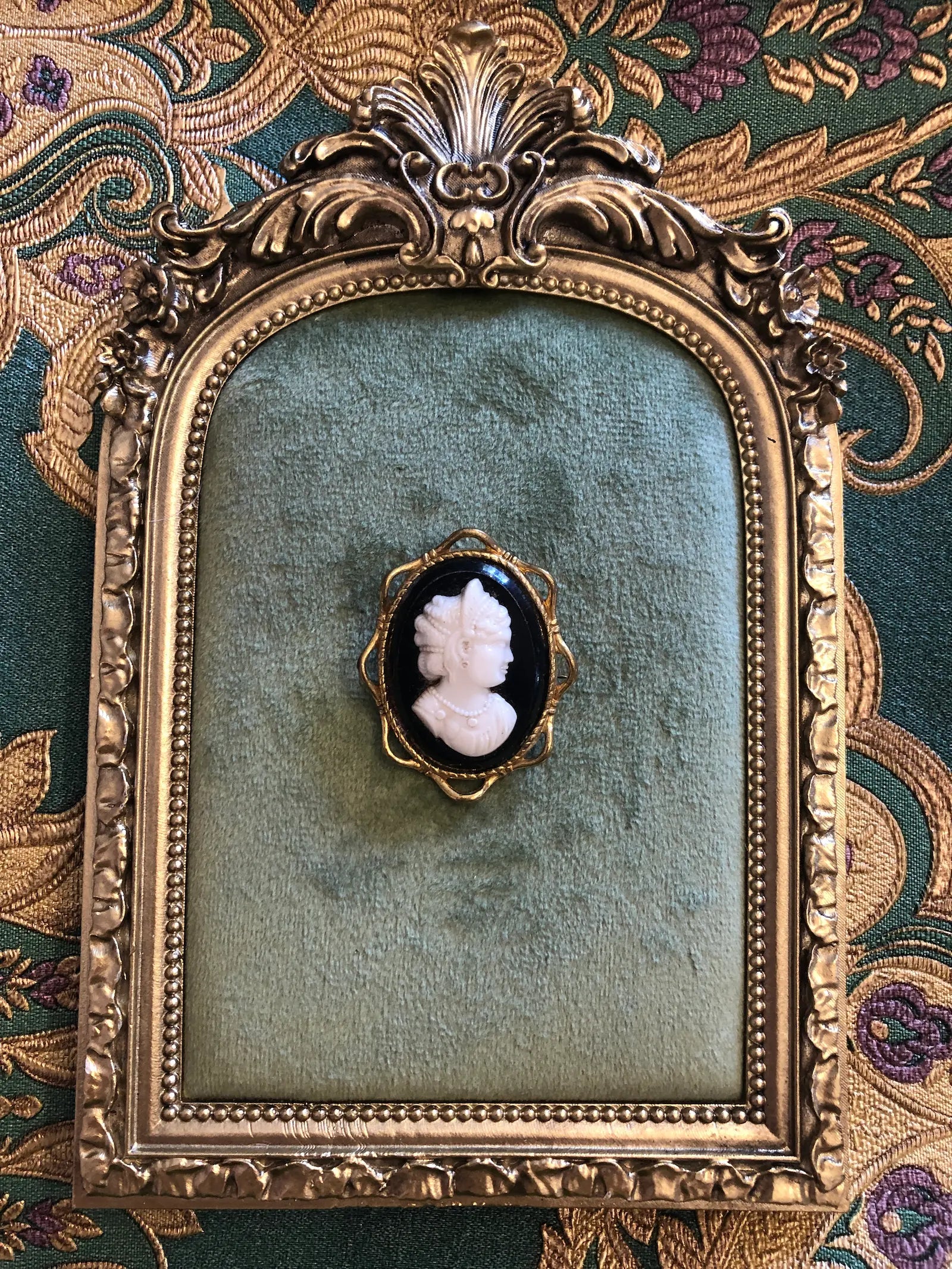 A Broochella Dea 4cm x 3cm vintage black and white cameo brooch featuring a carved white relief classical portrait of a noblewoman in profile with elaborate feathered and pearl upswept hair and a pearl necklace, set on a deep black oval ground within a gold-tone openwork filigree frame. On a green velvet tray with an ornate gold frame. The background is green and gold damask wallpaper