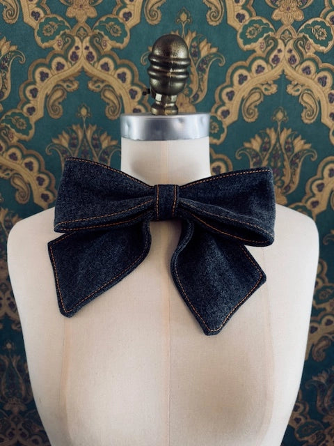 Broochella handmade medium denim upcycled jeans bow brooch. Pictured on a mannequin. The background is green and gold damask wallpaper.