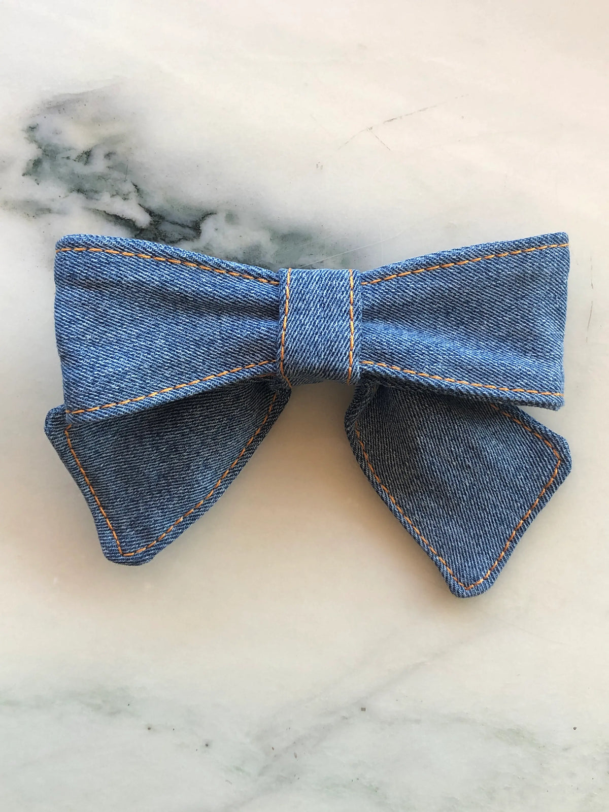 Broochella small sky blue denim bow brooch pin with contrast tan stitching, photographed on a white marble background.