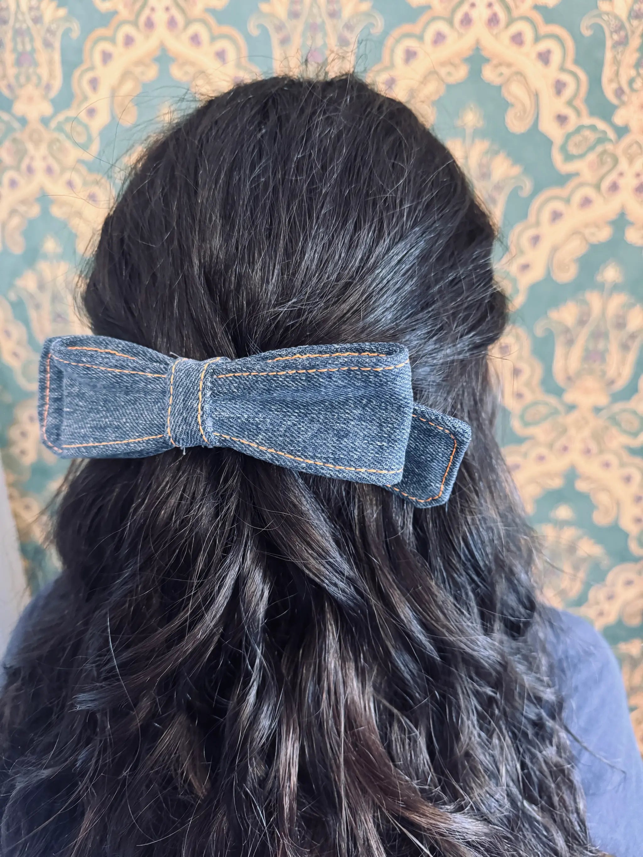 Broochella blue denim hair bow made from up cycled jeans. Pictured on a mannequin. The background is green and gold damask wallpaper.