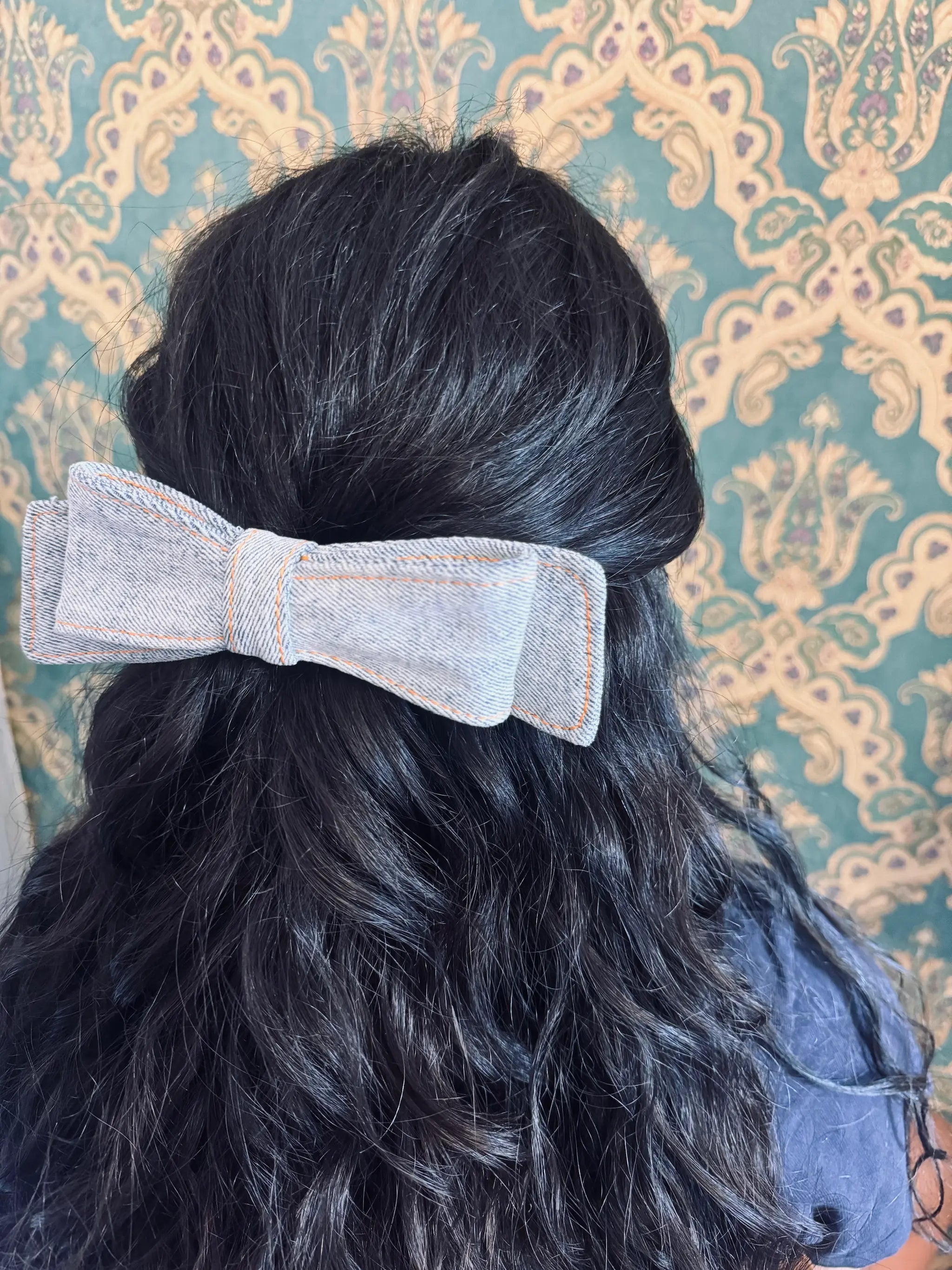 Broochella blue denim hair bow made from up cycled jeans. Pictured on a mannequin. The background is green and gold damask wallpaper.