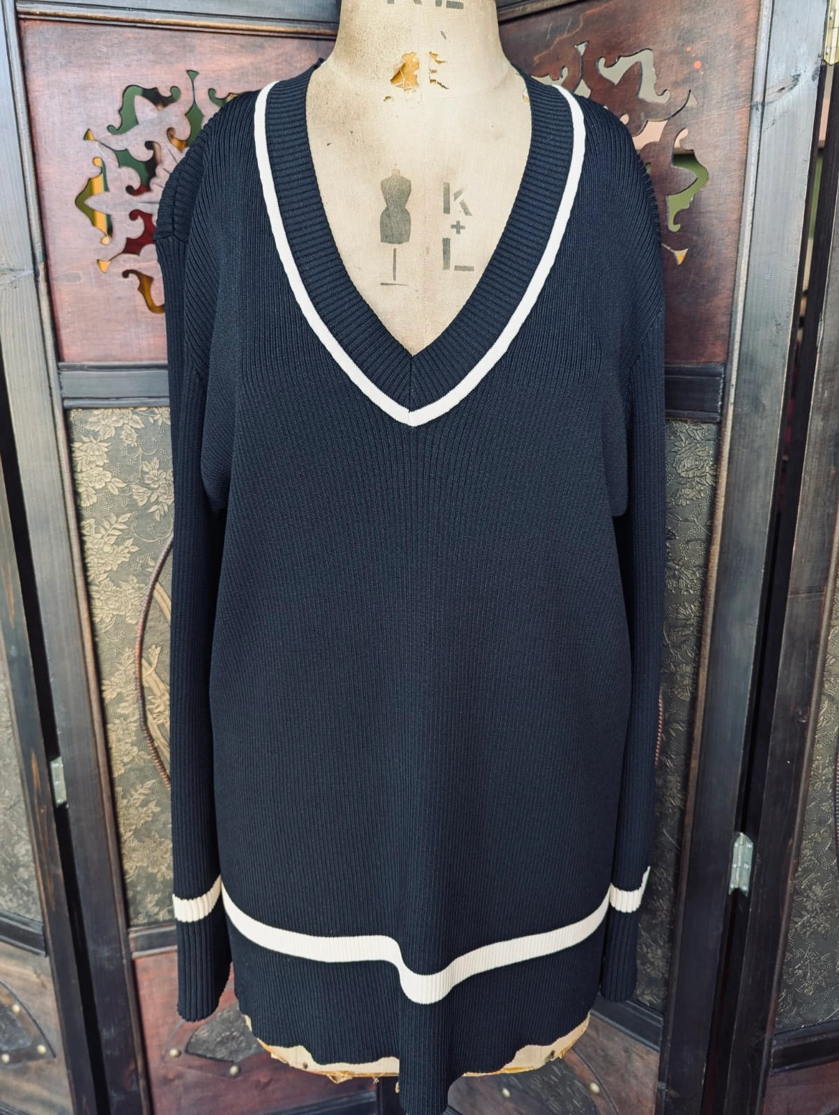 A Broochella Dissh black ribbed knit sweater with white contrast trim on the V-neck and hem, displayed on a dress form mannequin in front of a decorative wooden screen.