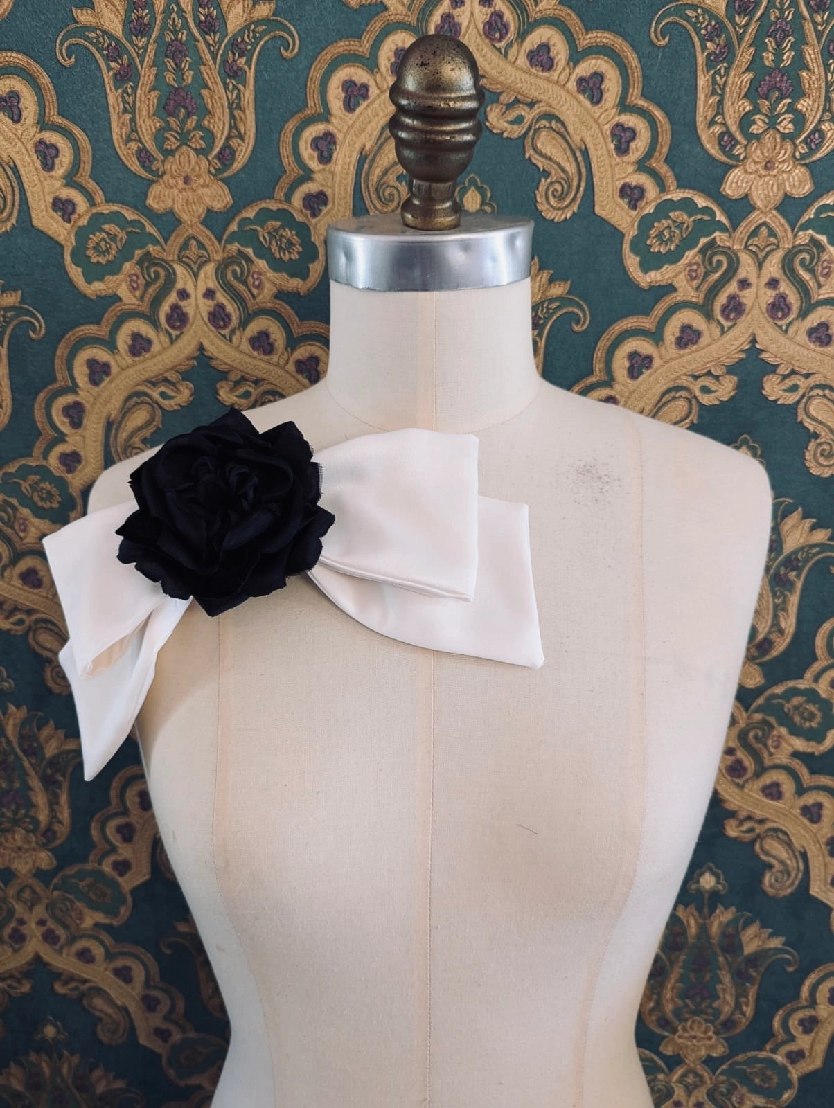 Broochella viva l'amor handmade ivory satin bow brooch with black flower centre.  It is pinned to a mannequin. The background is green and gold damask wallpaper.