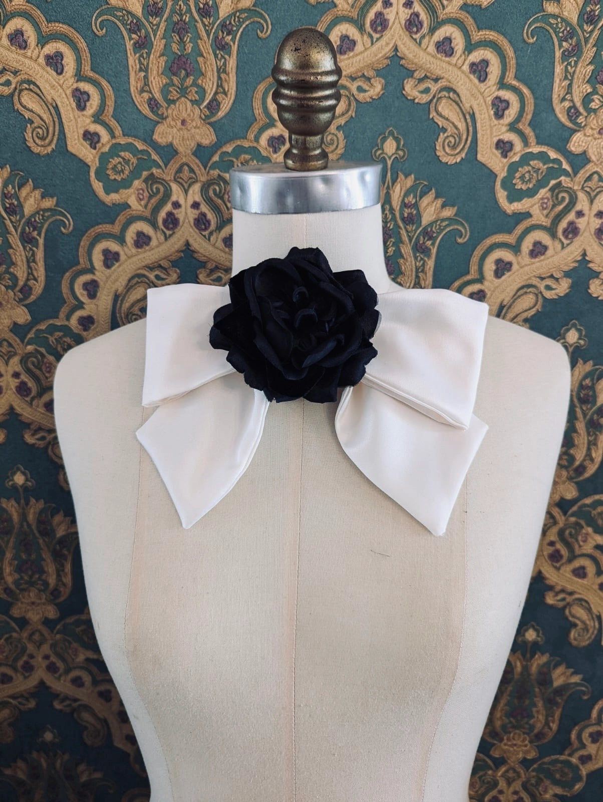 Broochella viva l'amor handmade ivory satin bow brooch with black flower centre.  It is pinned to a mannequin. The background is green and gold damask wallpaper.