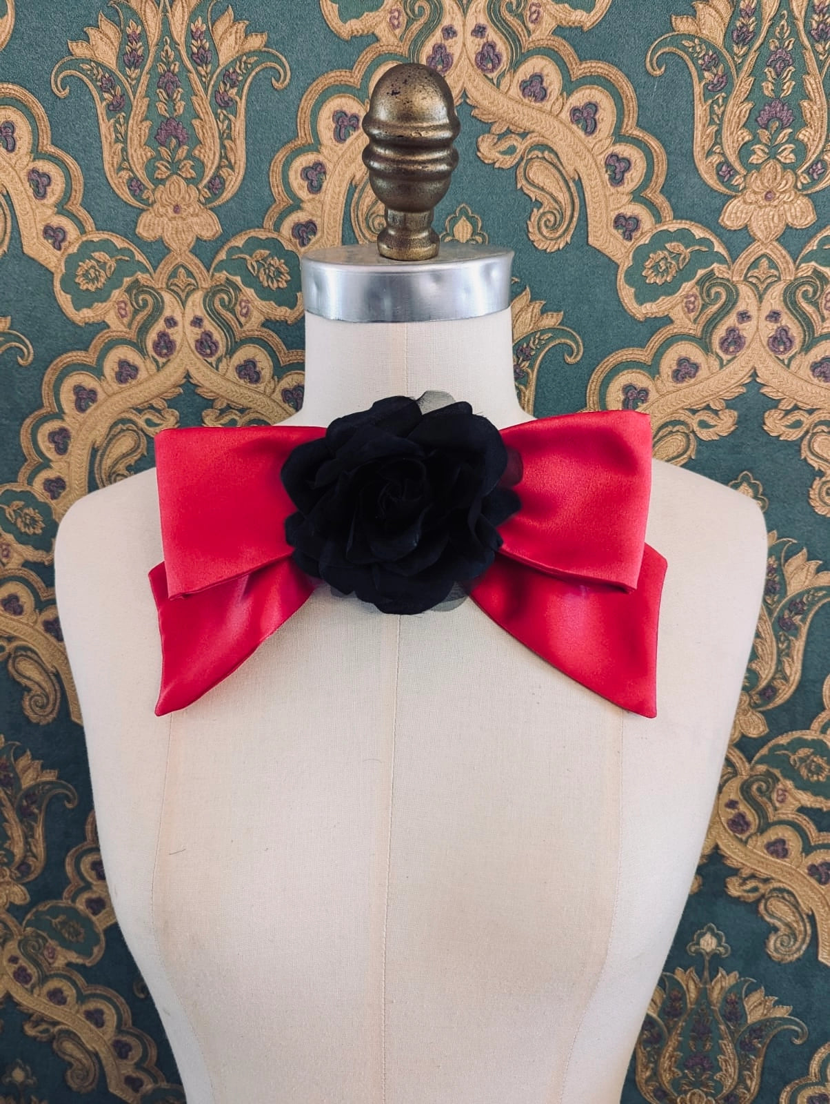 Broochella viva l'amor handmade pink satin bow brooch with black flower centre.  It is pinned to a mannequin. The background is green and gold damask wallpaper.