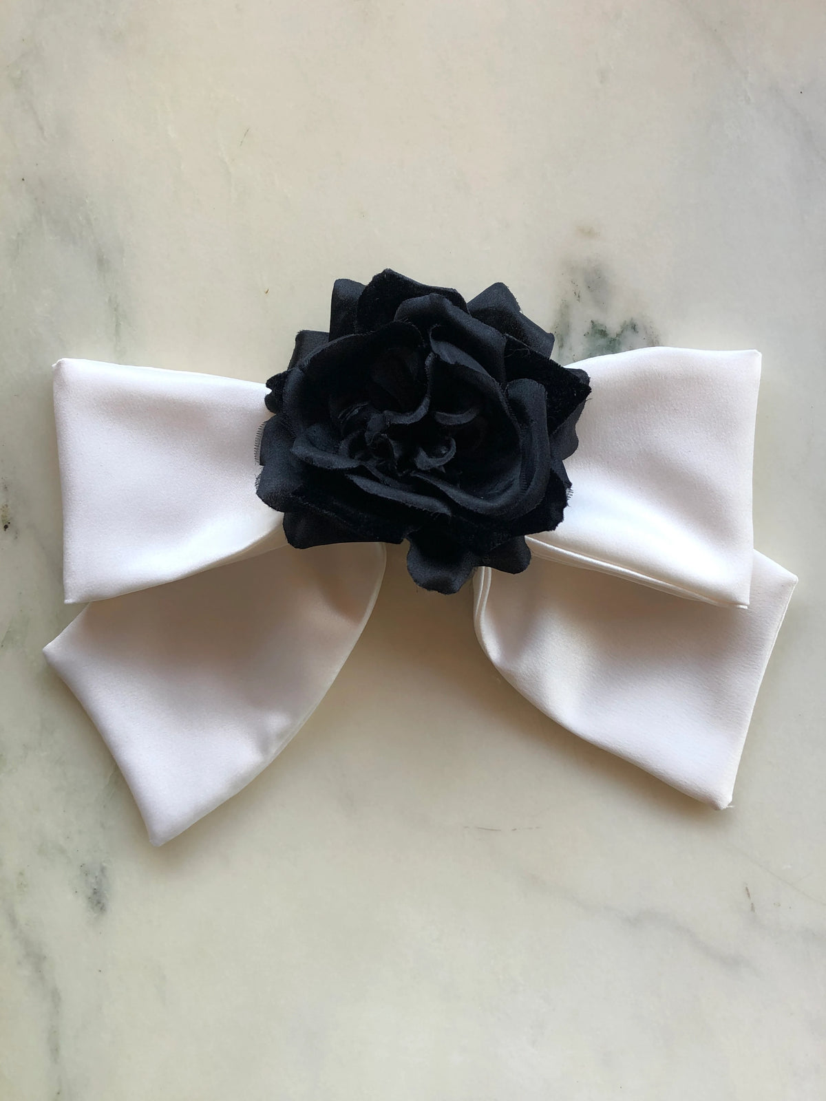 Broochella Dolce Amore Bridal Bow Brooch featuring an ivory satin statement bow with a sculptural black flower at the centre, photographed on a pale cream marble background.